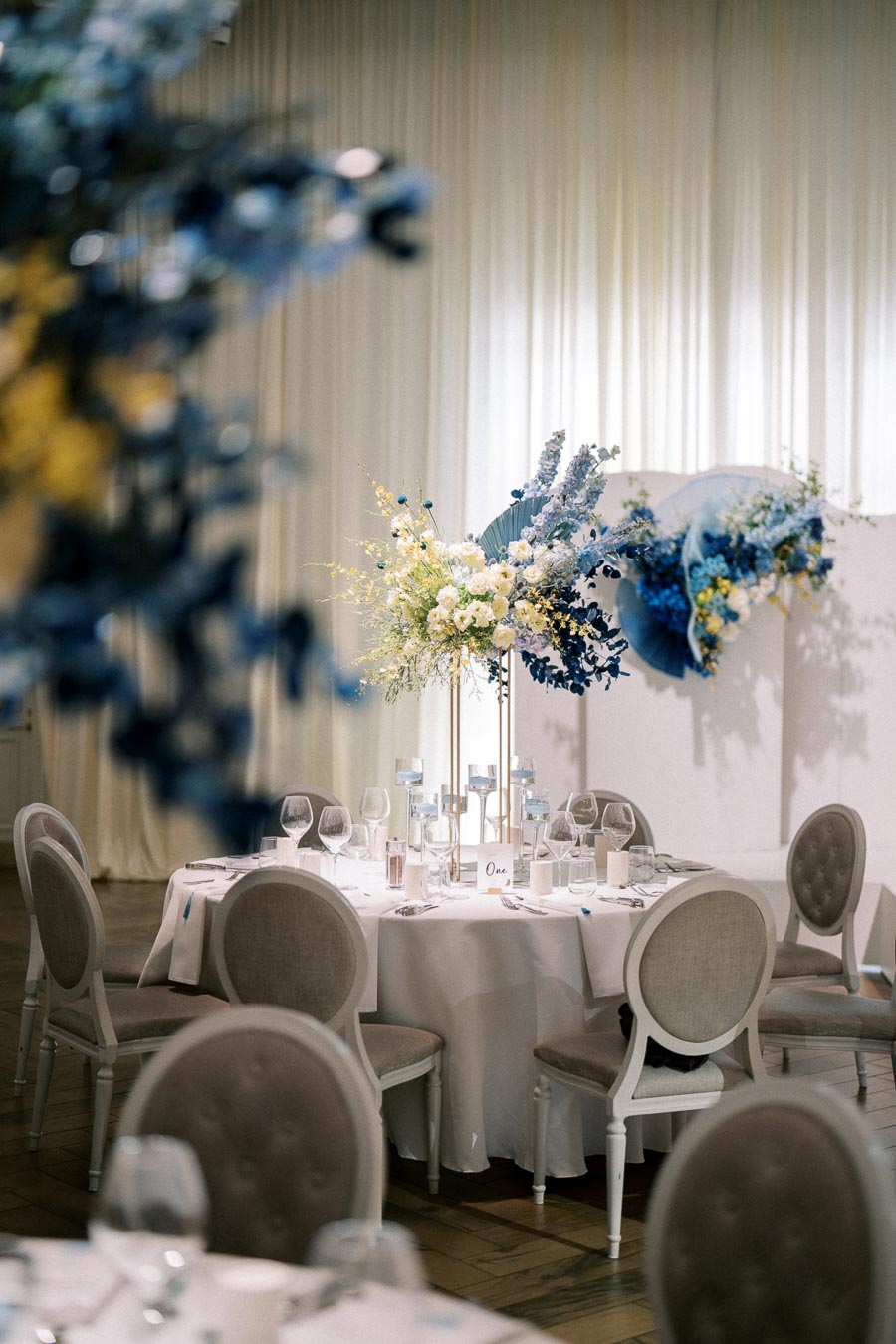 Elegant wedding reception table setup with white tablecloth, fine china, wine glasses, and a centerpiece featuring tall floral arrangements in shades of blue and white. The soft drapery in the background adds to the sophisticated ambiance of the event space.