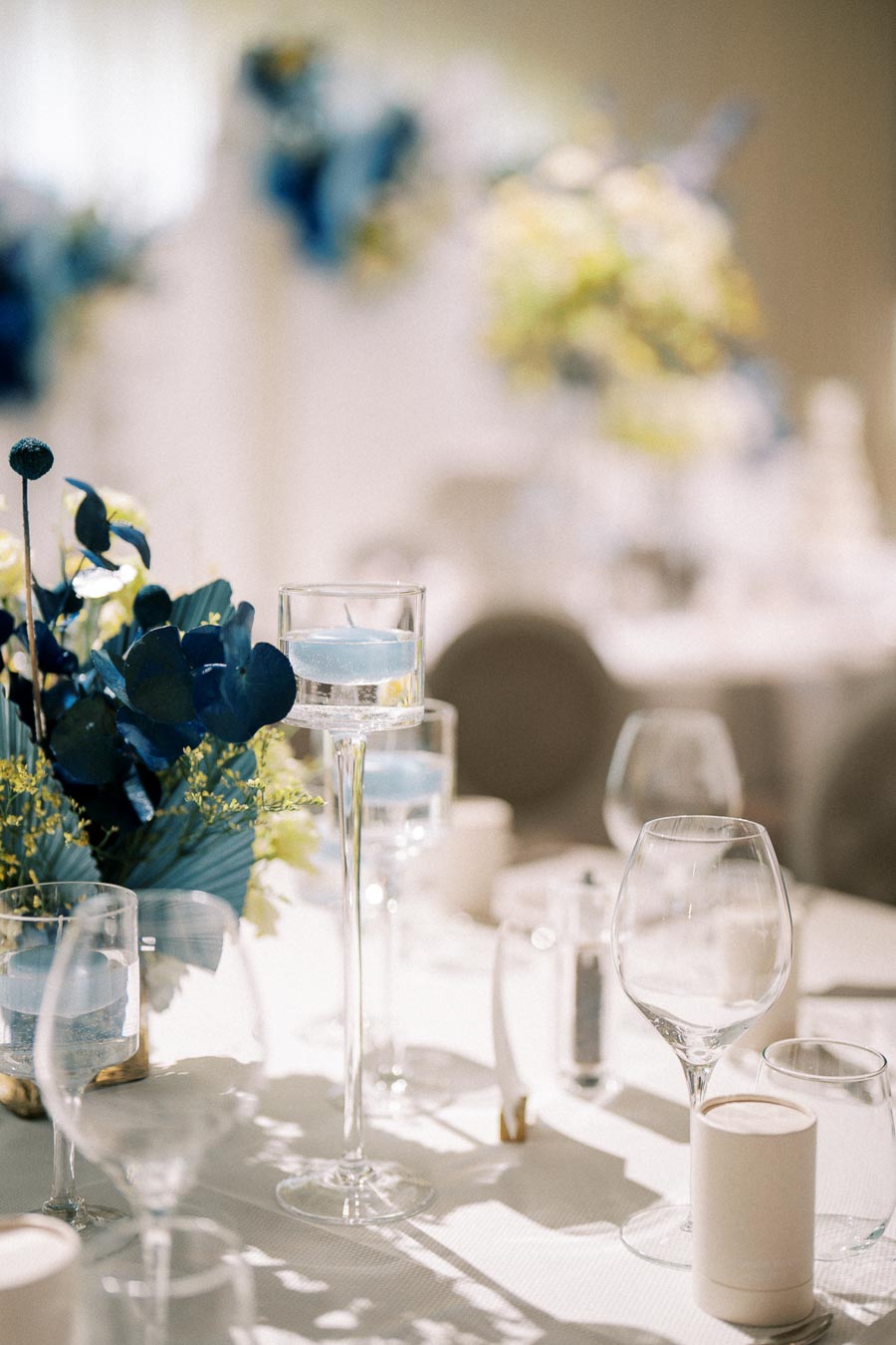Elegant wedding table setting with blue floral centerpiece, tall glass candle holders, and pristine glassware on a white tablecloth.