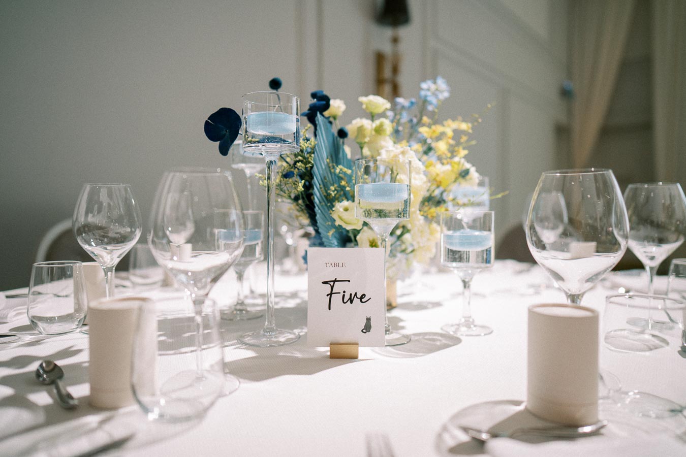 Elegant wedding table setup with Table Five sign, featuring blue and white floral centerpiece, tall candle holders with blue candles, and neatly arranged glassware on a white tablecloth.