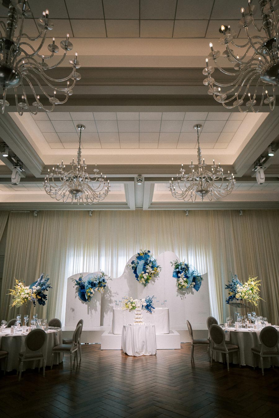 Elegant wedding reception venue with floral decorations in blue and white, featuring round tables set with glassware, a stylish wooden floor, and grand chandeliers overhead.
