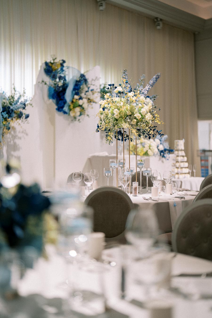Elegant wedding reception setup with tables decorated with tall floral centerpieces featuring white and blue flowers, surrounded by neatly arranged glassware and tableware in a softly lit venue.