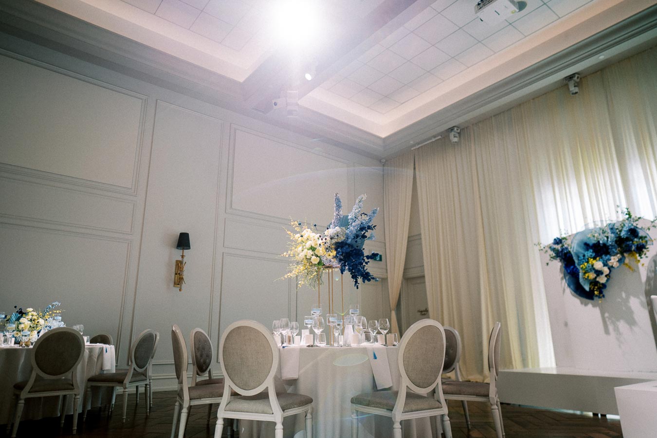 Elegant ballroom wedding reception with white round tables, chic chairs, and stunning blue and white floral centerpieces against cream drapery and a high ceiling, perfect for a sophisticated event setting.