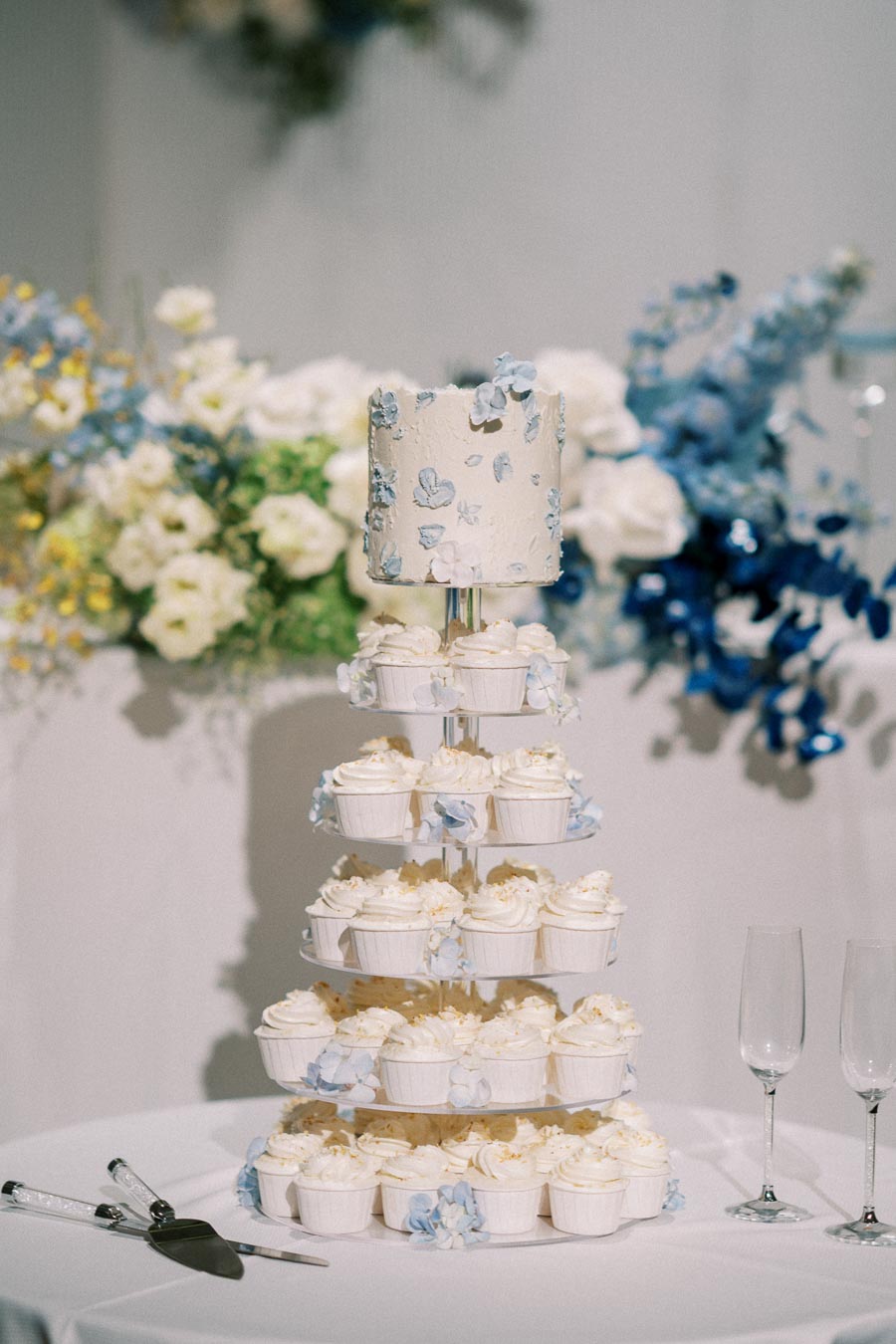 Elegant tiered wedding cupcake display with a floral-decorated mini cake on top, surrounded by delicate white and blue flowers, set on a white tablecloth with serving utensils and champagne glasses.