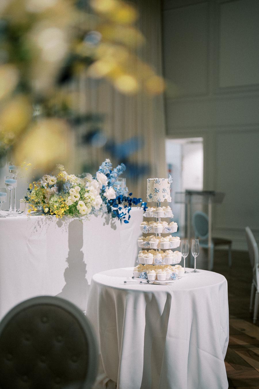 Elegant wedding reception setup featuring a tiered cake stand with cupcakes and a small cake on top, surrounded by floral arrangements in shades of white and blue, and two champagne glasses on a round white table.