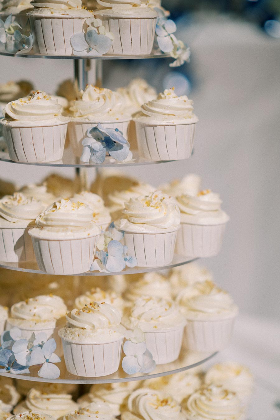 Three-tiered stand of elegant white cupcakes with creamy frosting and delicate blue hydrangea decorations, perfect for a wedding or special celebration.