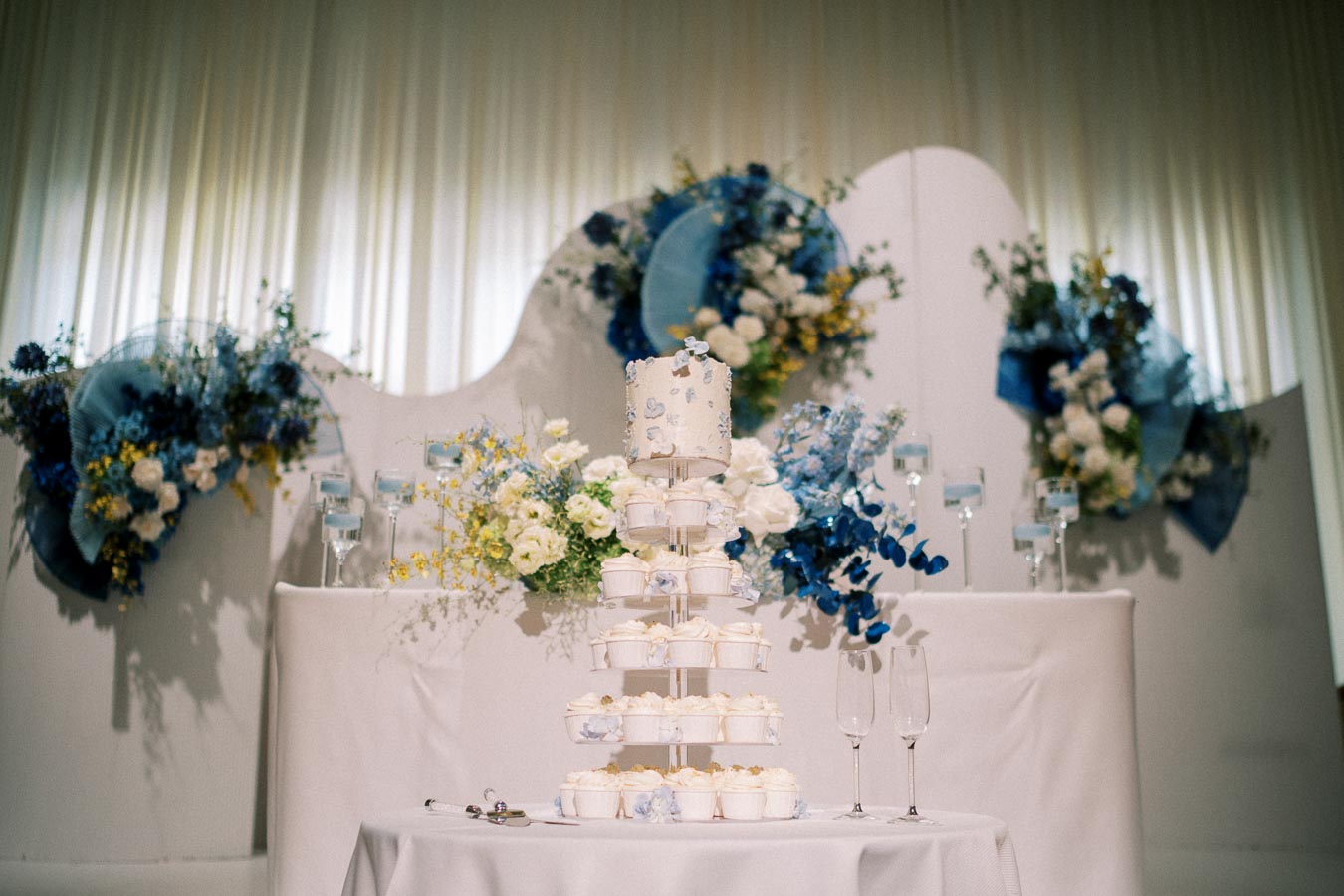 Elegant wedding cake display with white and blue floral decorations, featuring a tiered cake surrounded by cupcakes and champagne glasses, set against a sophisticated backdrop.