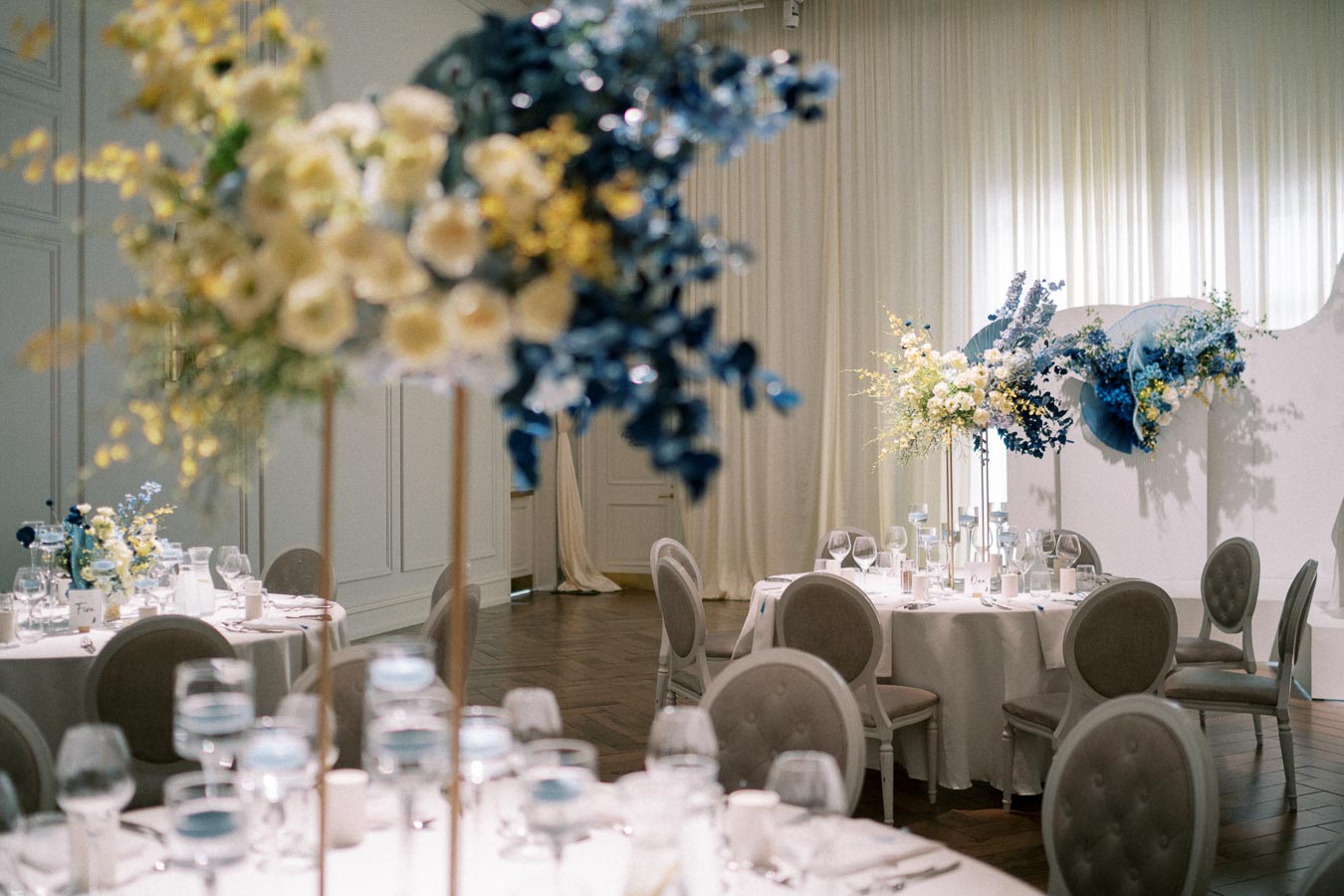 Elegant wedding reception setup with round tables adorned with blue and yellow floral arrangements, glassware, and candles, set in a softly lit banquet hall with light curtains and wooden floors.