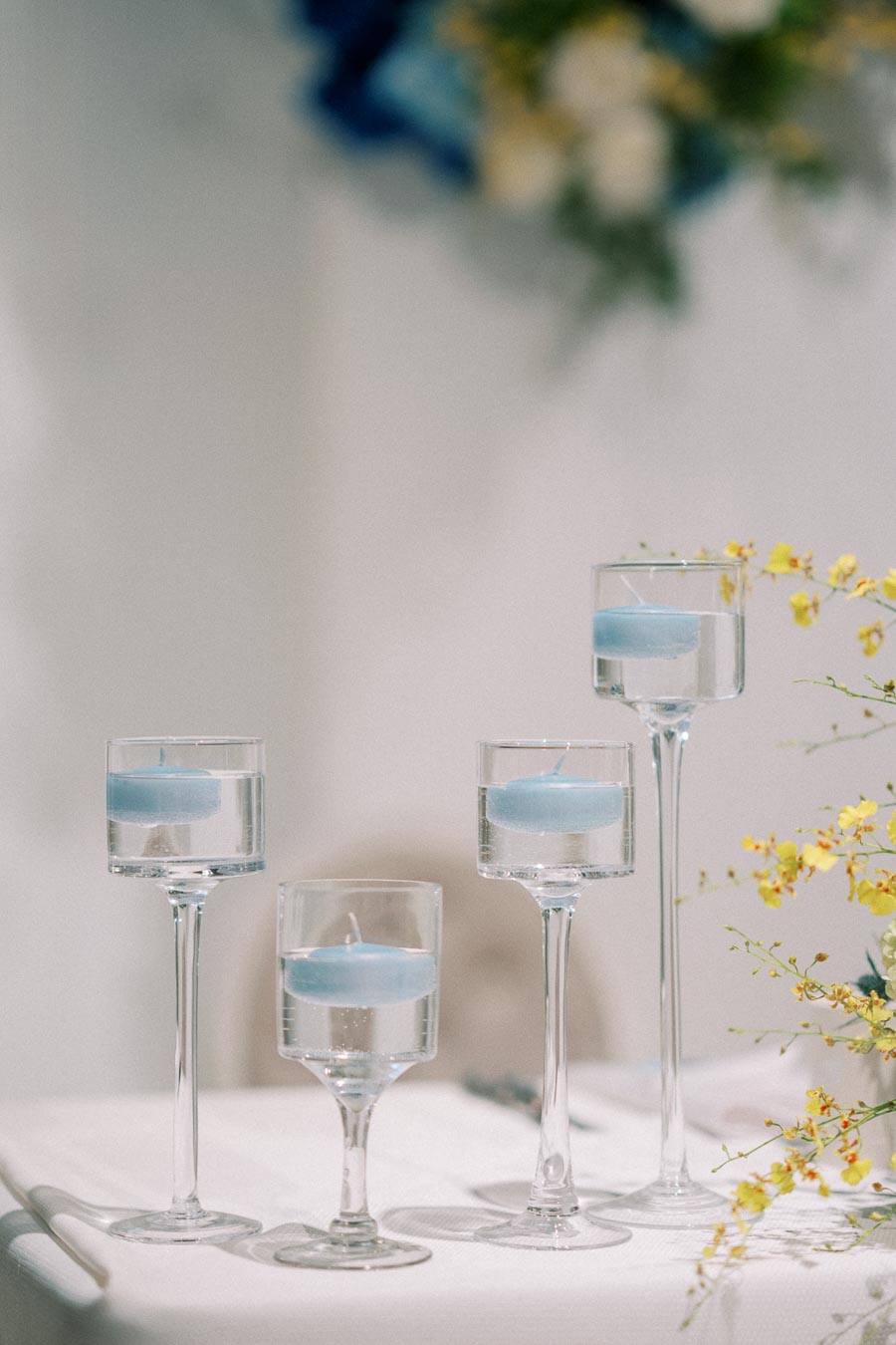 Elegant table setting with tall glass holders and floating blue candles, surrounded by delicate yellow flowers. Perfect for a sophisticated wedding or romantic dinner atmosphere.