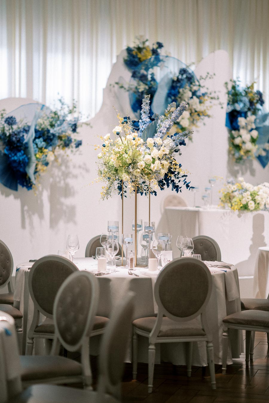 Elegant wedding reception setup featuring round tables adorned with white tablecloths, beige cushioned chairs, and tall floral centerpieces. Blue and white floral arrangements decorate the background, creating a sophisticated and inviting atmosphere.