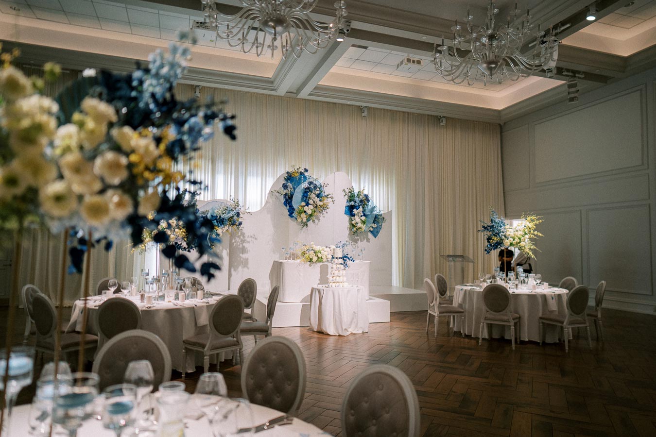 Elegant wedding reception venue with a beautifully decorated room featuring round tables adorned with white linens and floral centerpieces in blue and white hues, alongside a multi-tiered wedding cake displayed on a pedestal, surrounded by ambient lighting and chandeliers.
