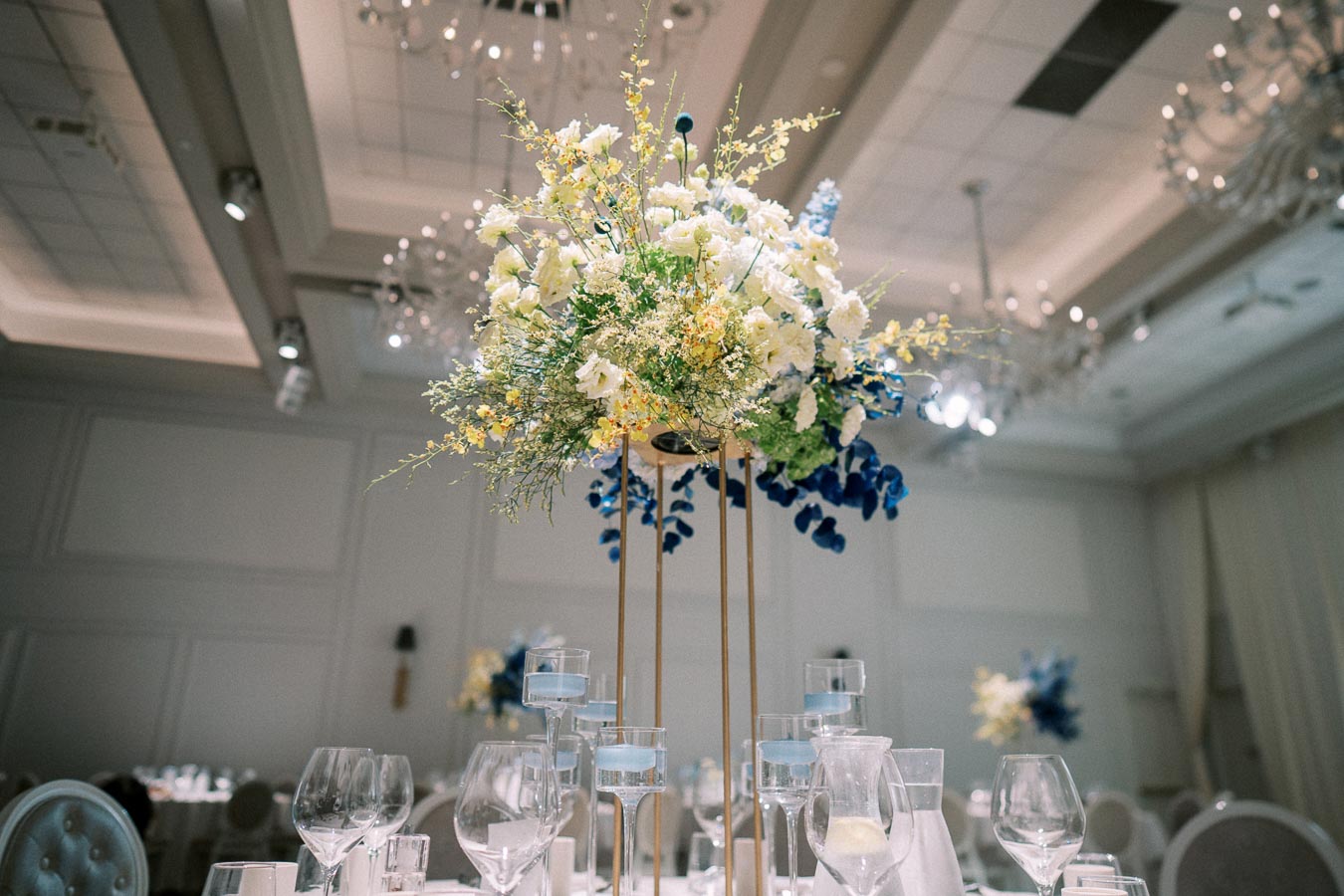 Elegant wedding reception table with tall floral centerpiece featuring white, yellow, and blue flowers, surrounded by glassware and decorated with votive candles in a luxurious banquet hall setting.
