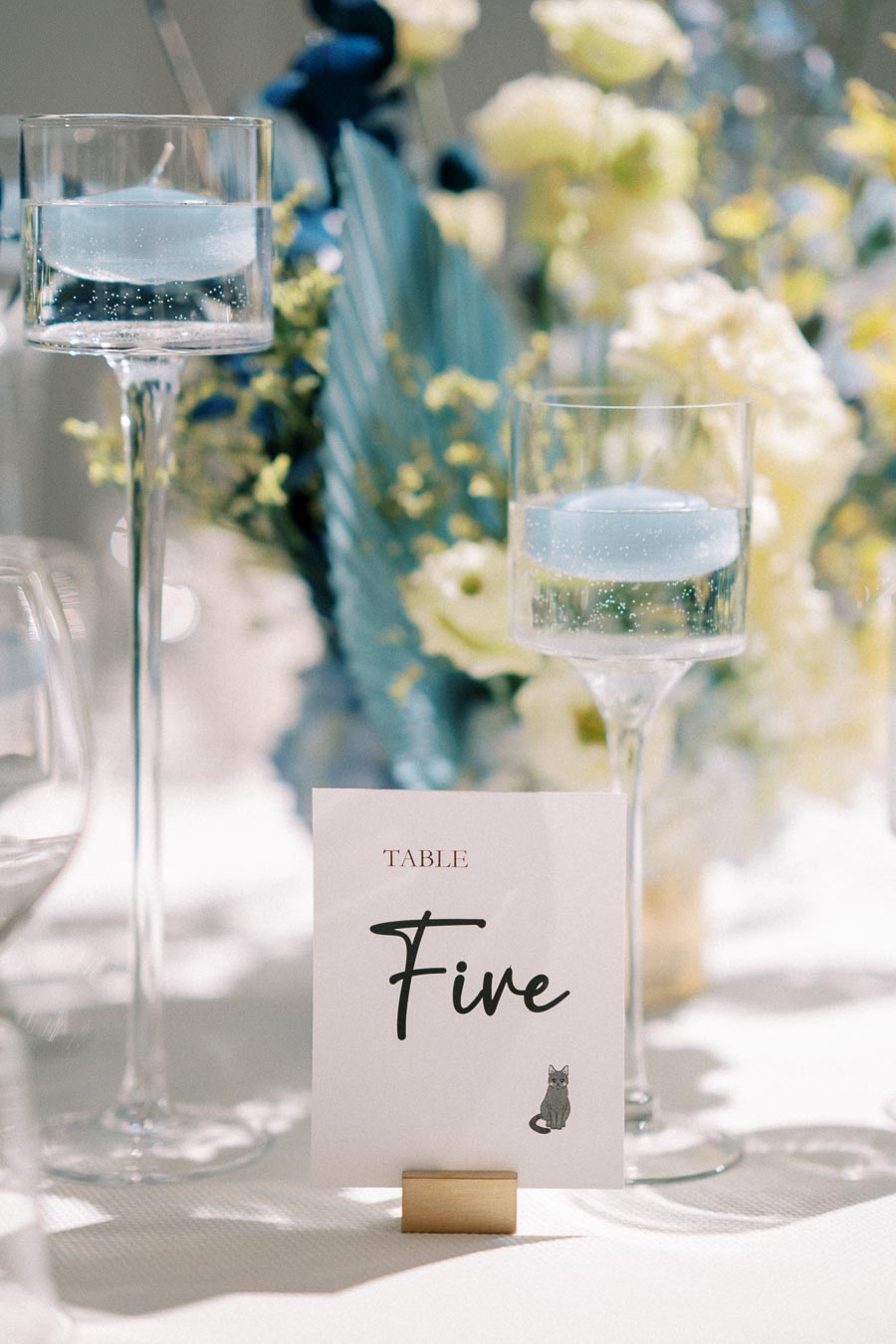 Elegant wedding table setting with tall glass candle holders and a table card labeled Table Five, accompanied by blue and yellow floral arrangements, creating a refined and romantic atmosphere.