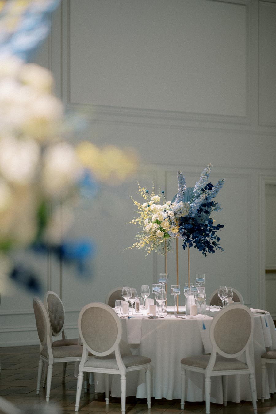 Elegant wedding table setting with white tablecloth, surrounded by stylish white chairs. The centerpiece features tall blue and white floral arrangements, creating a sophisticated and romantic atmosphere. Perfect decor inspiration for weddings and formal events.