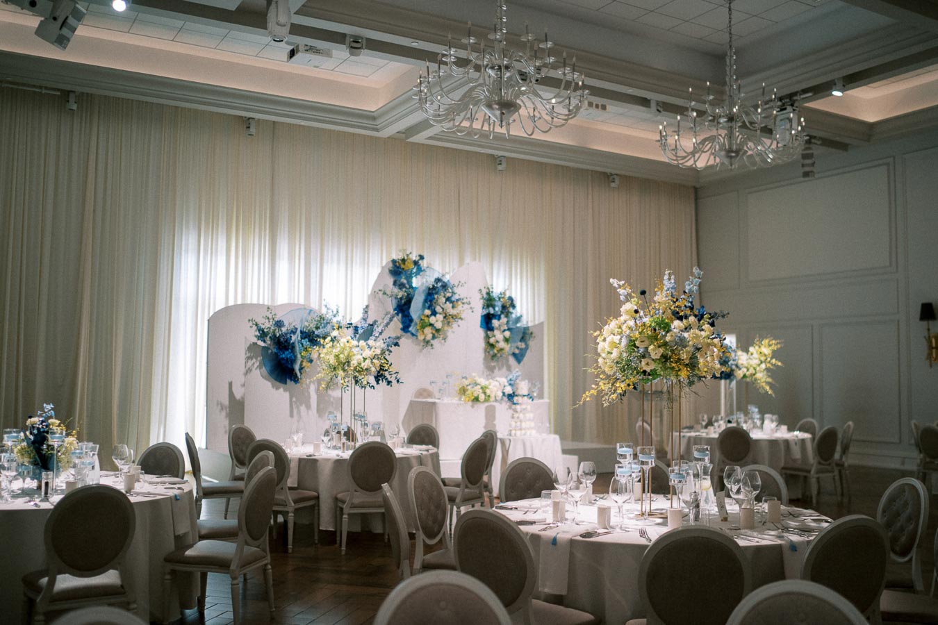 Elegant wedding reception hall with round tables, white tablecloths, and neutral chairs. The decor includes large floral arrangements in shades of blue and yellow, set against a backdrop of soft drapery and white wall panels, under ornate chandeliers.