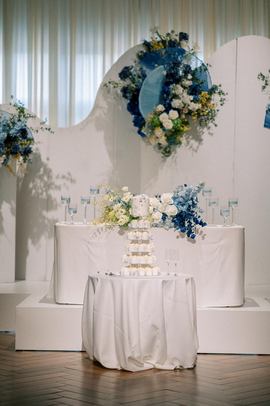 Elegant wedding dessert table with white and blue floral arrangements, tiered cake, and wine glasses, set in a sophisticated reception setting.