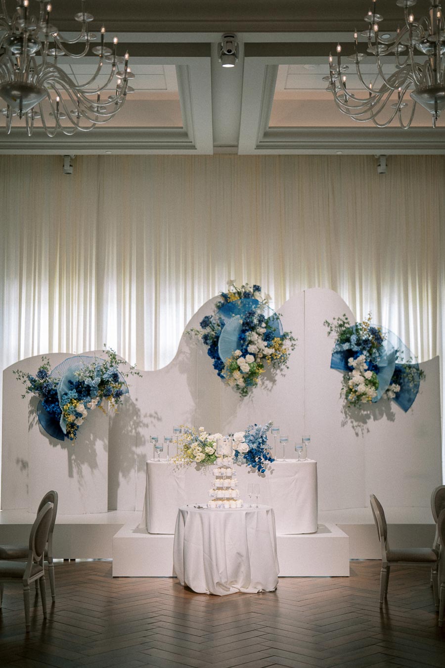 Elegant wedding venue setup featuring a white stage adorned with blue and white floral arrangements, a tiered cake on a table, and chandeliers hanging from the ceiling.
