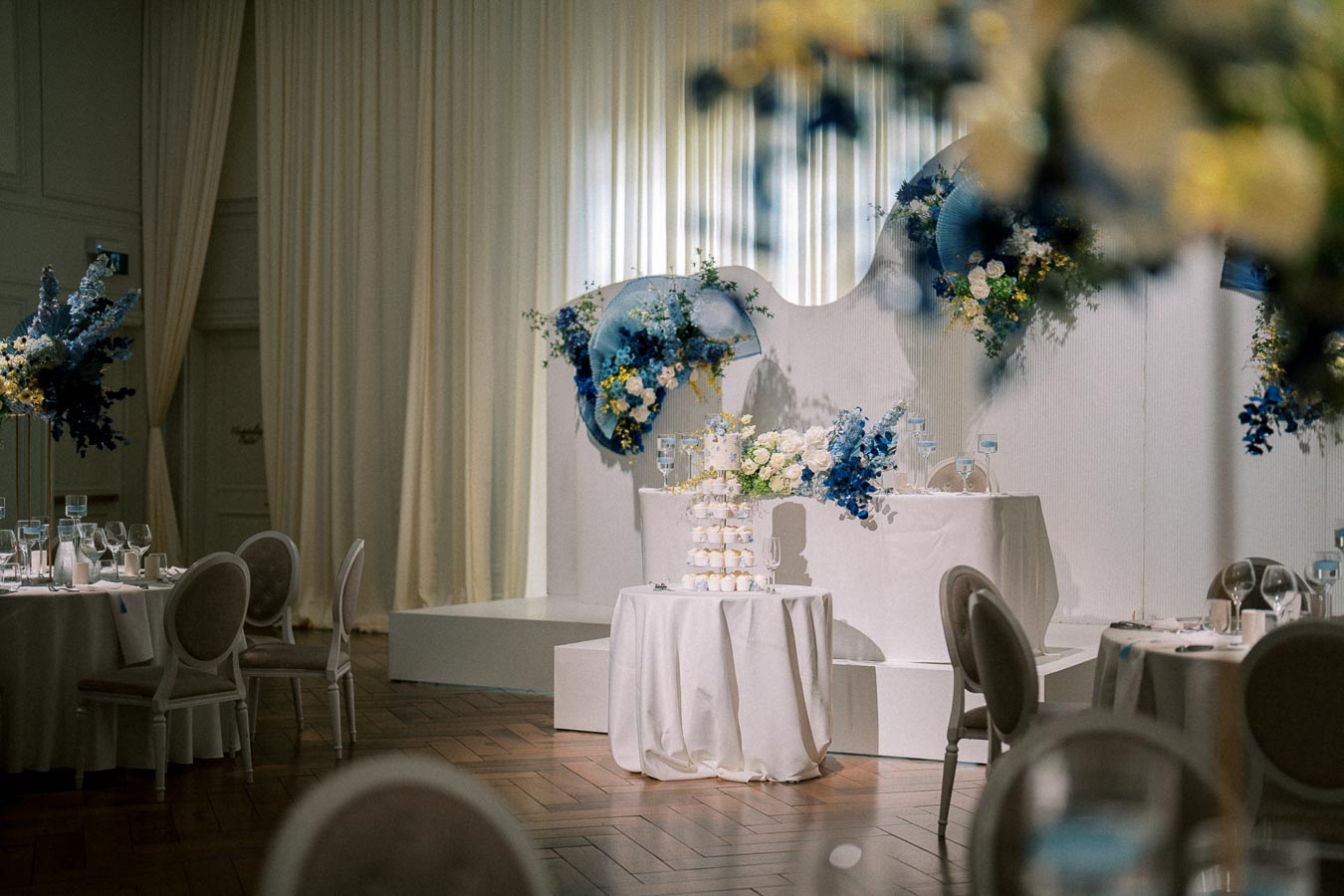 Elegant wedding reception setup featuring beautifully arranged tables with white linens and neutral chairs, decorated with white and blue floral arrangements, and a multi-tiered display of white desserts against a soft curtain backdrop.