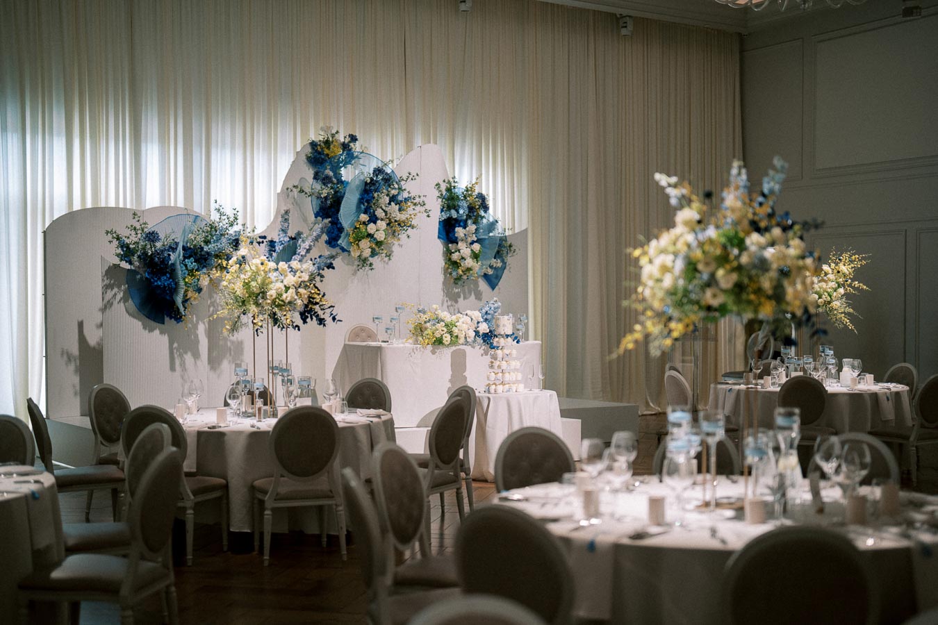 Elegant wedding reception setup with round tables adorned with white tablecloths and floral centerpieces, featuring blue and white flowers. The backdrop displays a beautiful floral arrangement with a soft beige curtain in the background, creating a sophisticated and celebratory atmosphere.
