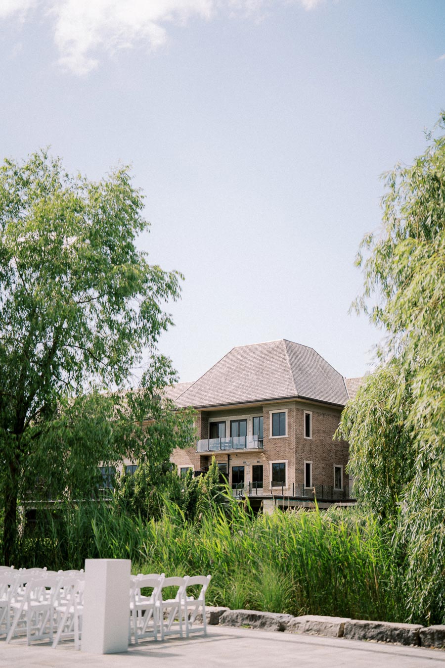 Brick building with a pitched roof surrounded by lush greenery and clear skies, featuring a setup of white chairs and a podium, suggesting an outdoor event or ceremony.