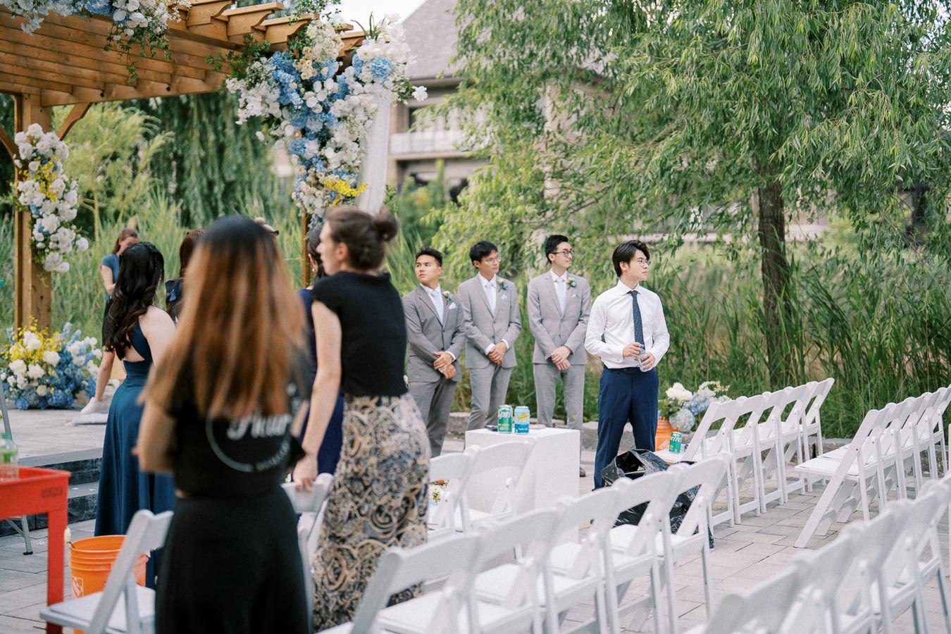 Outdoor wedding ceremony setup with floral arch, groomsmen in gray suits, and guests preparing.