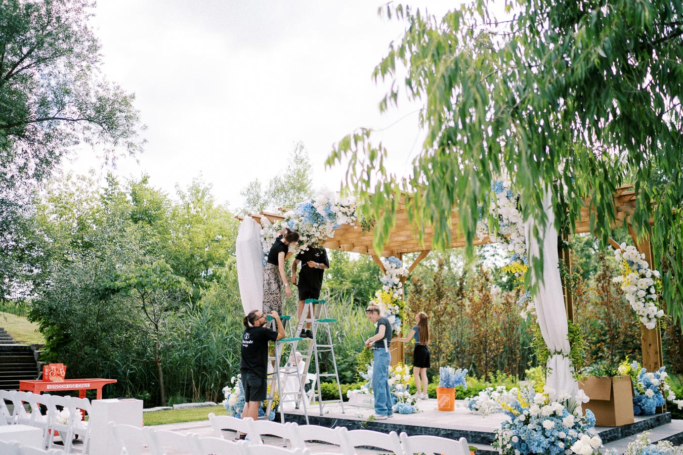 Outdoor wedding ceremony setup with workers arranging floral decorations on a wooden pergola. White and blue flowers adorn the structure, surrounded by lush greenery and white chairs in the foreground, creating an elegant garden wedding atmosphere.
