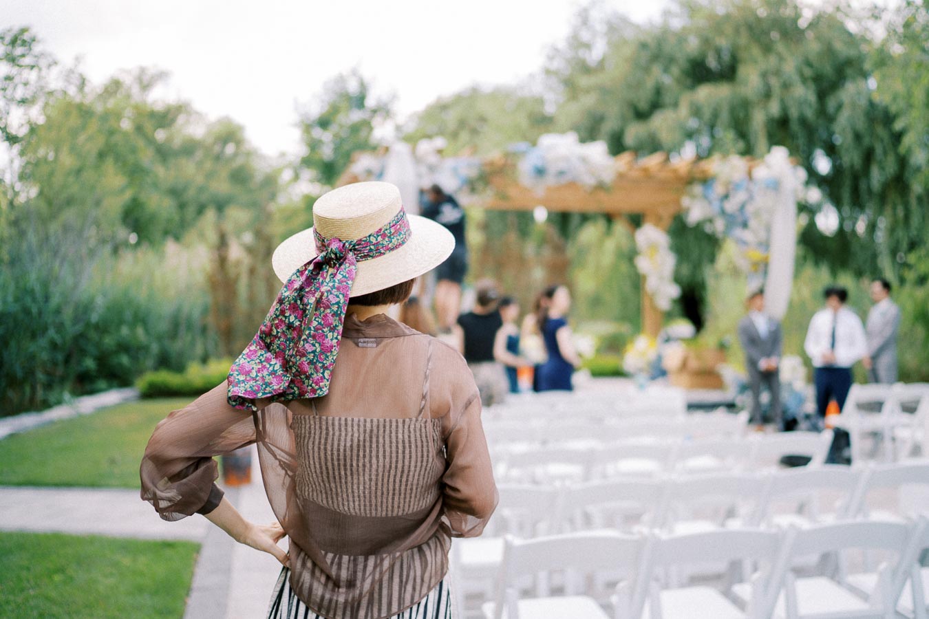 A woman wearing a straw hat with a floral scarf stands in a garden setting, observing a wedding ceremony setup with white chairs and a decorated wooden arbor in the background.