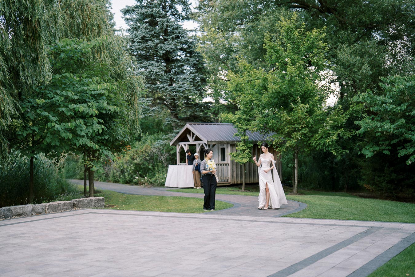 Wedding scene in a lush garden setting with a bride in a white gown walking along a pathway. A small wooden structure and other guests are visible in the background surrounded by green trees and grass.