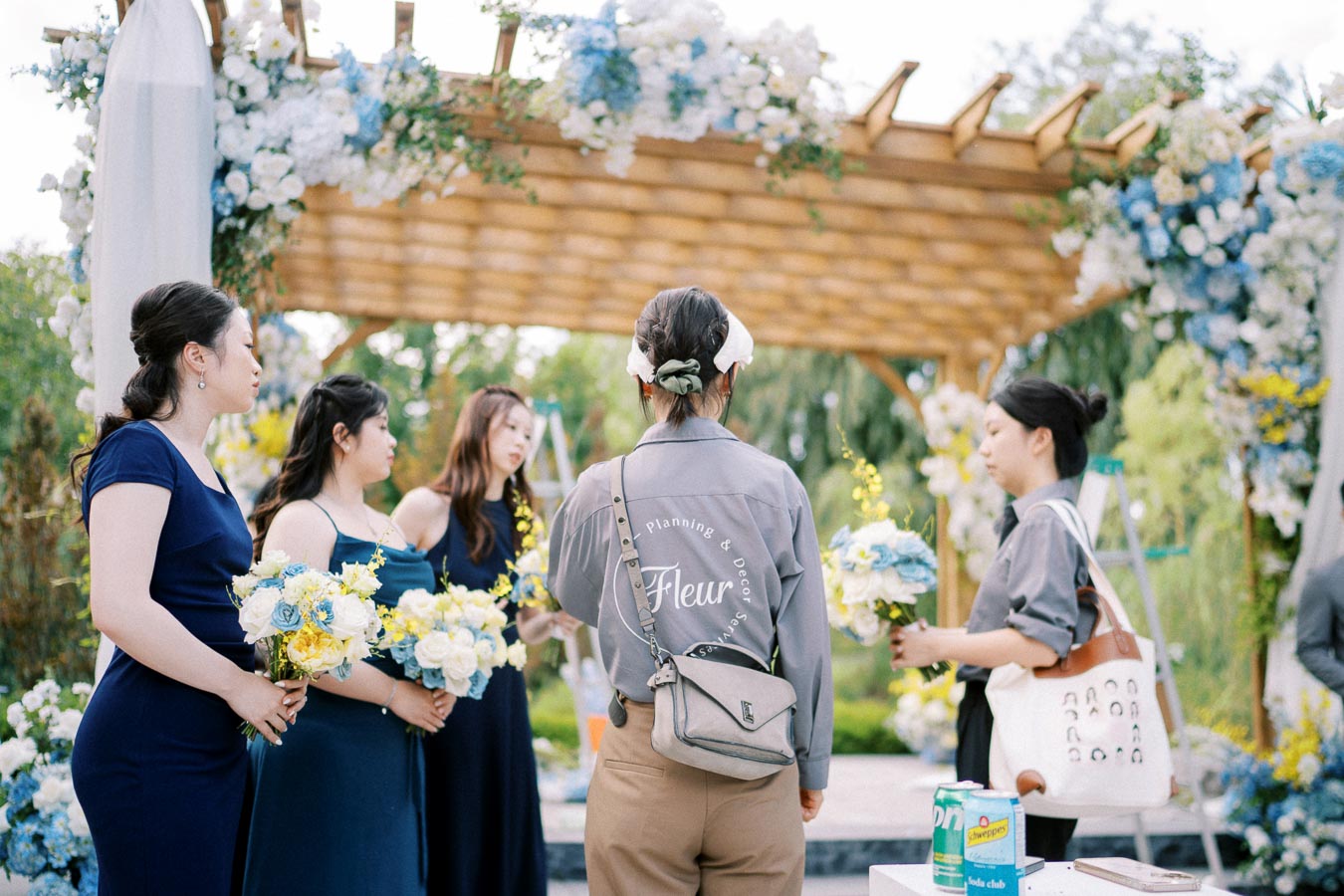 Wedding ceremony preparation with a florist team arranging bouquets under a floral arch, featuring blue and yellow flowers. Women in blue dresses stand nearby, adding final touches. The outdoor setting is lush with greenery.