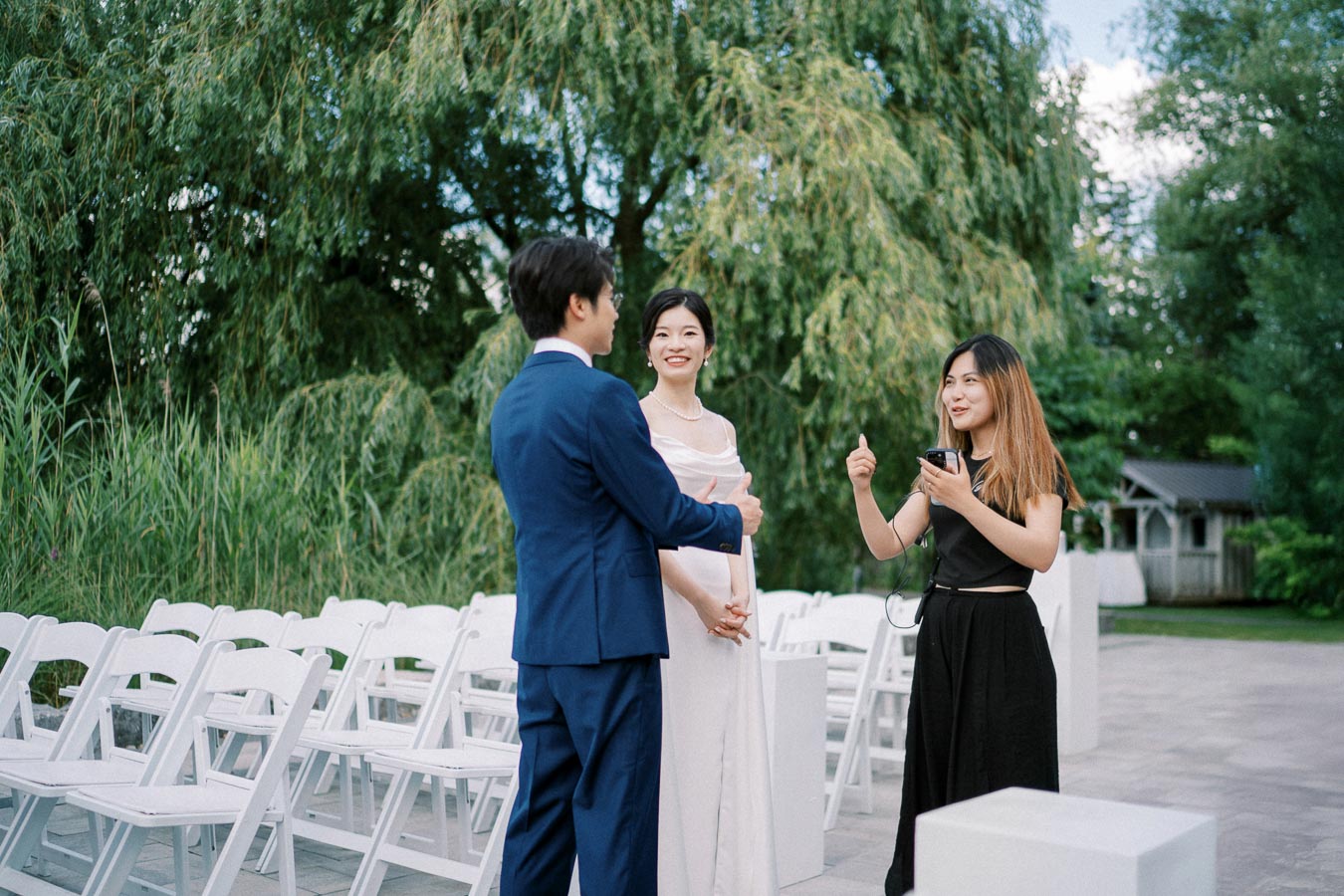 A couple stands smiling at an outdoor wedding ceremony, with a woman holding a microphone and giving a thumbs up, surrounded by greenery and rows of white chairs.