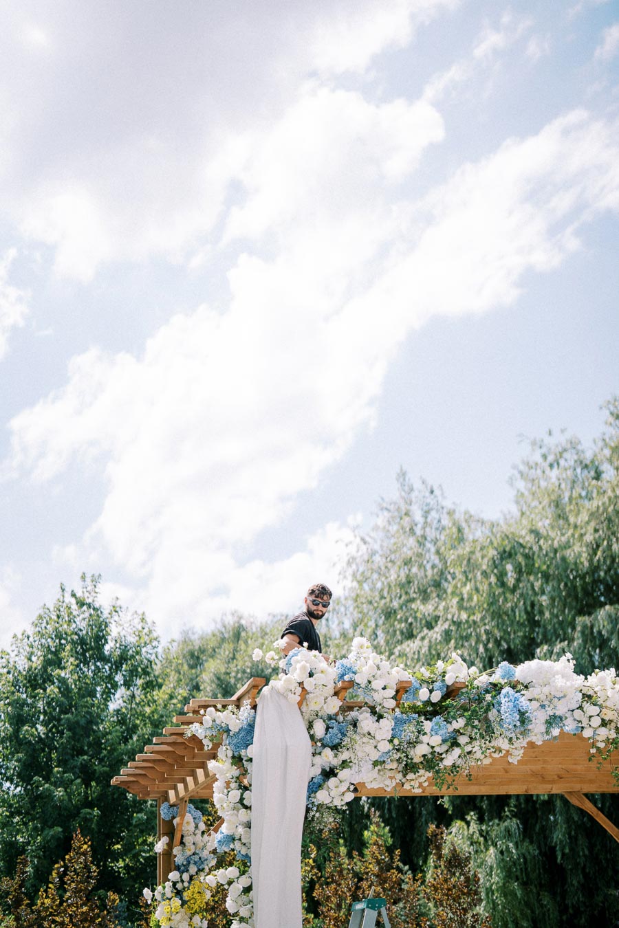 A person decorates a wooden pergola with an arrangement of white and blue flowers against a backdrop of a bright sky and lush greenery.