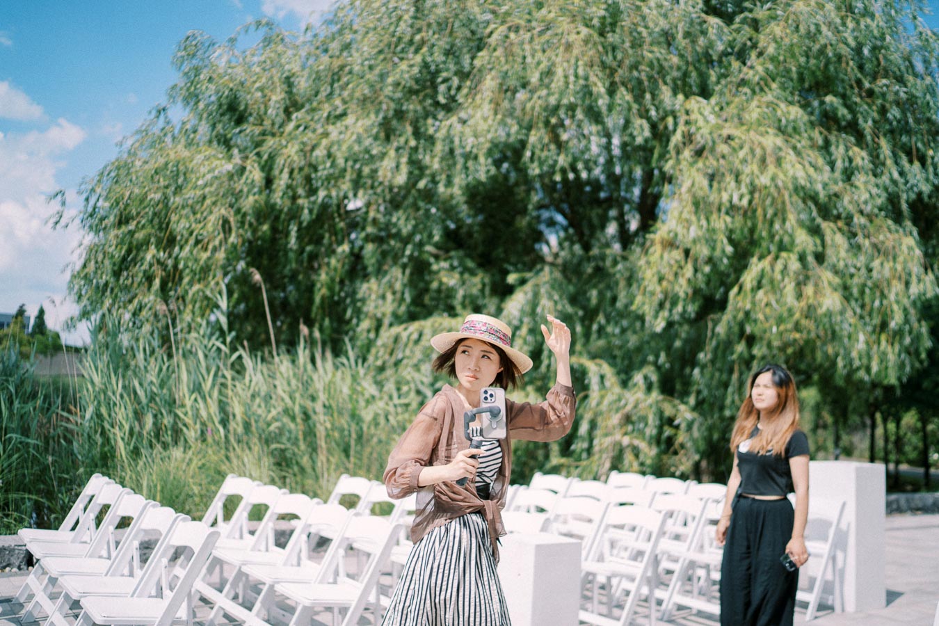 A woman in a striped dress and straw hat holding a smartphone walks through an outdoor setting with rows of white folding chairs, surrounded by lush green trees under a bright blue sky.