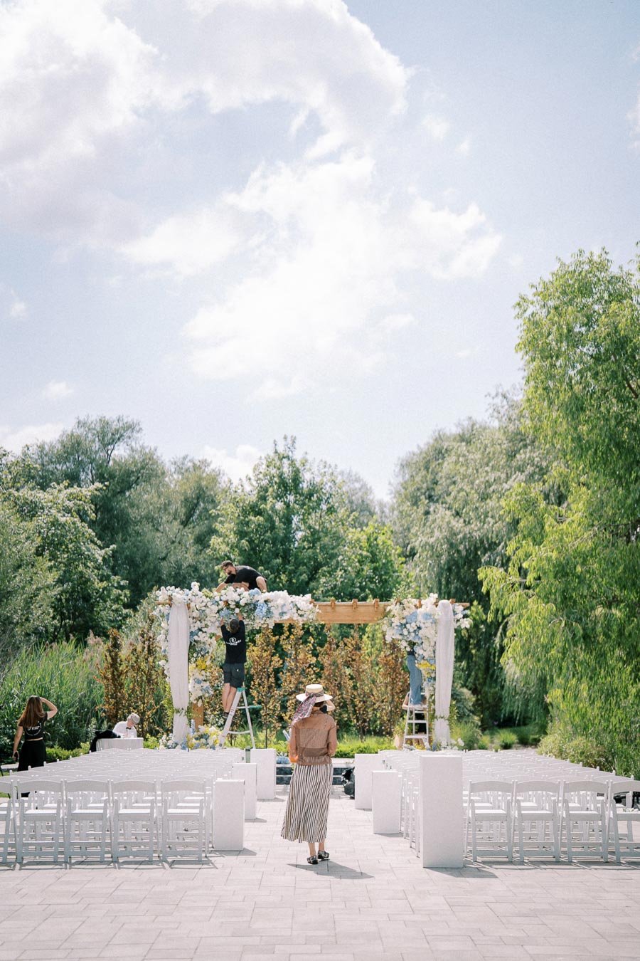 Outdoor wedding setup in progress with floral arch and white chairs under a sunny sky surrounded by greenery.