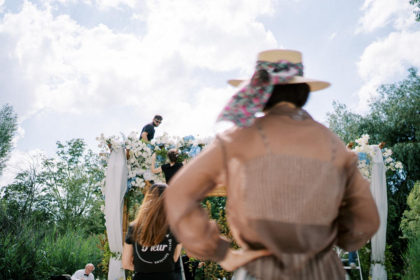 Outdoor wedding setup in progress, featuring a floral arch being decorated by event staff under a sunny sky, with a woman in a hat observing the scene.