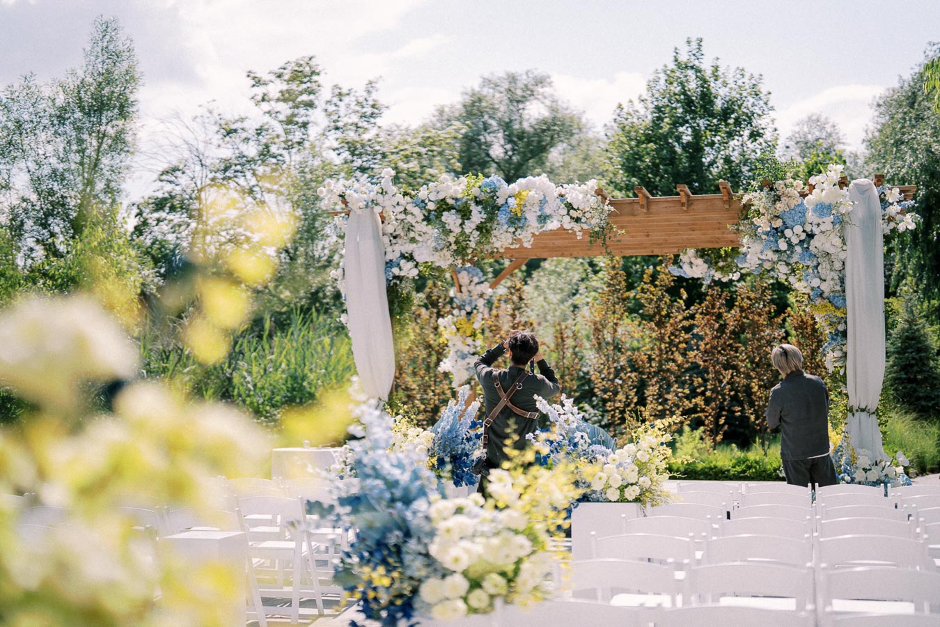 Outdoor wedding ceremony setup with a beautifully decorated wooden arch adorned with white and blue flowers and flowing fabric, surrounded by lush greenery and rows of white chairs.