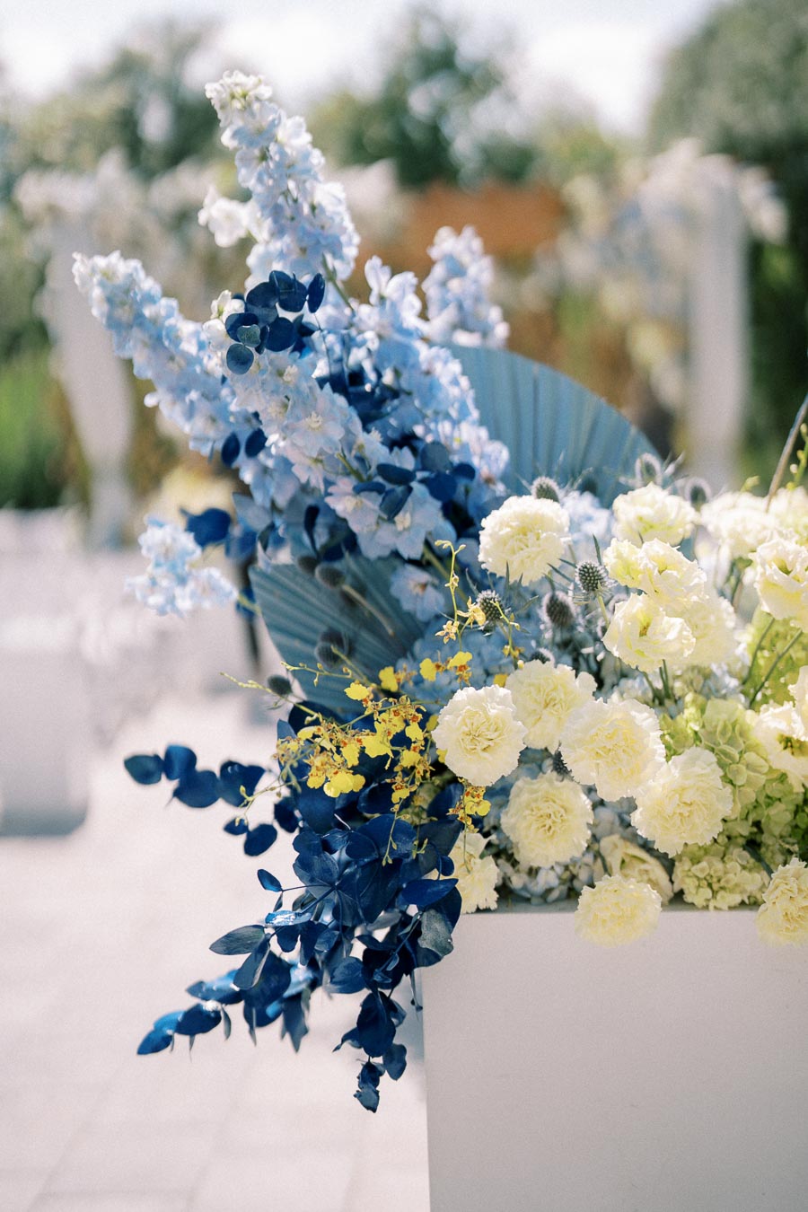 Elegant floral arrangement with blue delphiniums, yellow orchids, and cream carnations in a white vase, set against a blurred garden background.