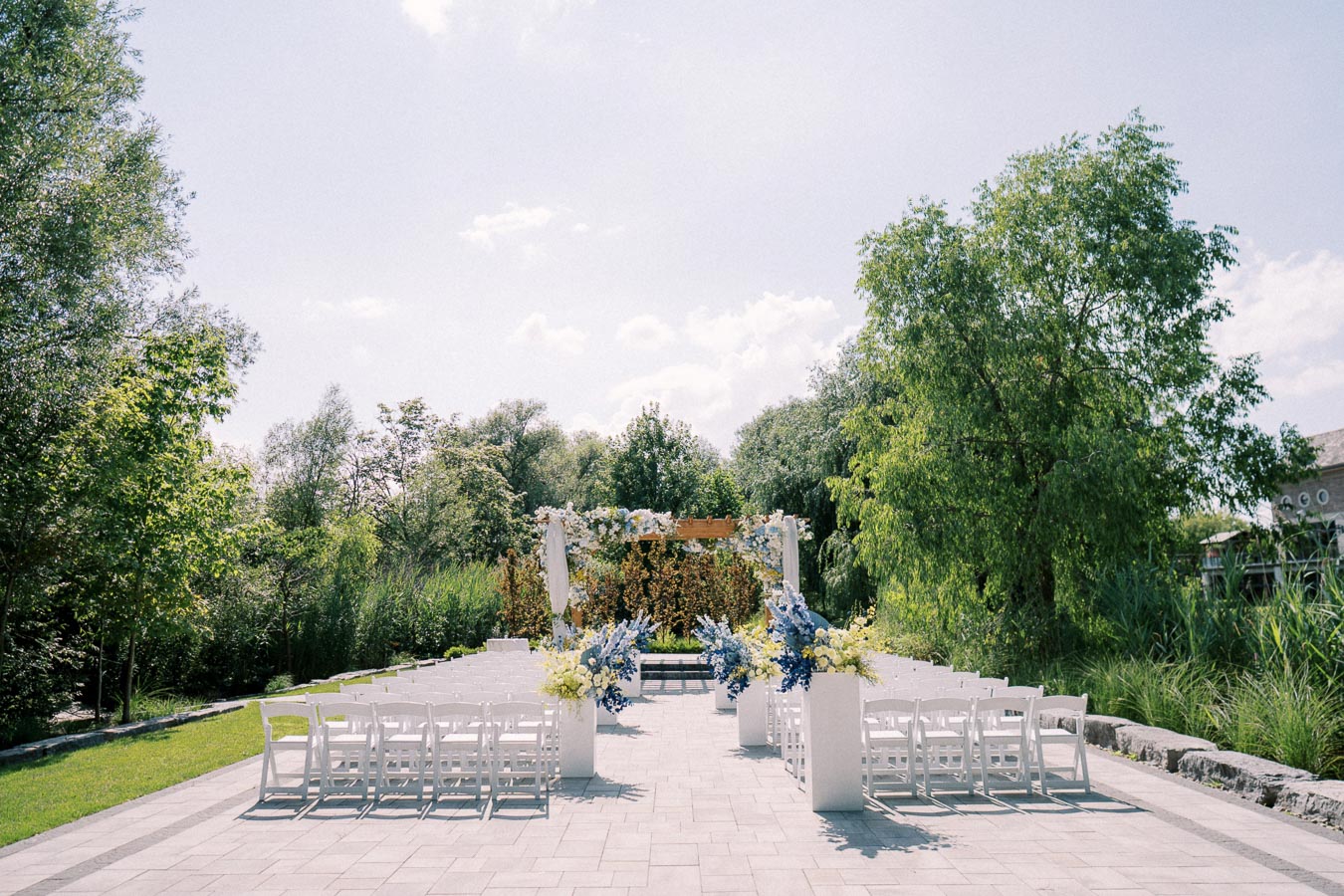 Outdoor wedding ceremony setup with white chairs and floral decorations under a pergola in a garden on a sunny day.