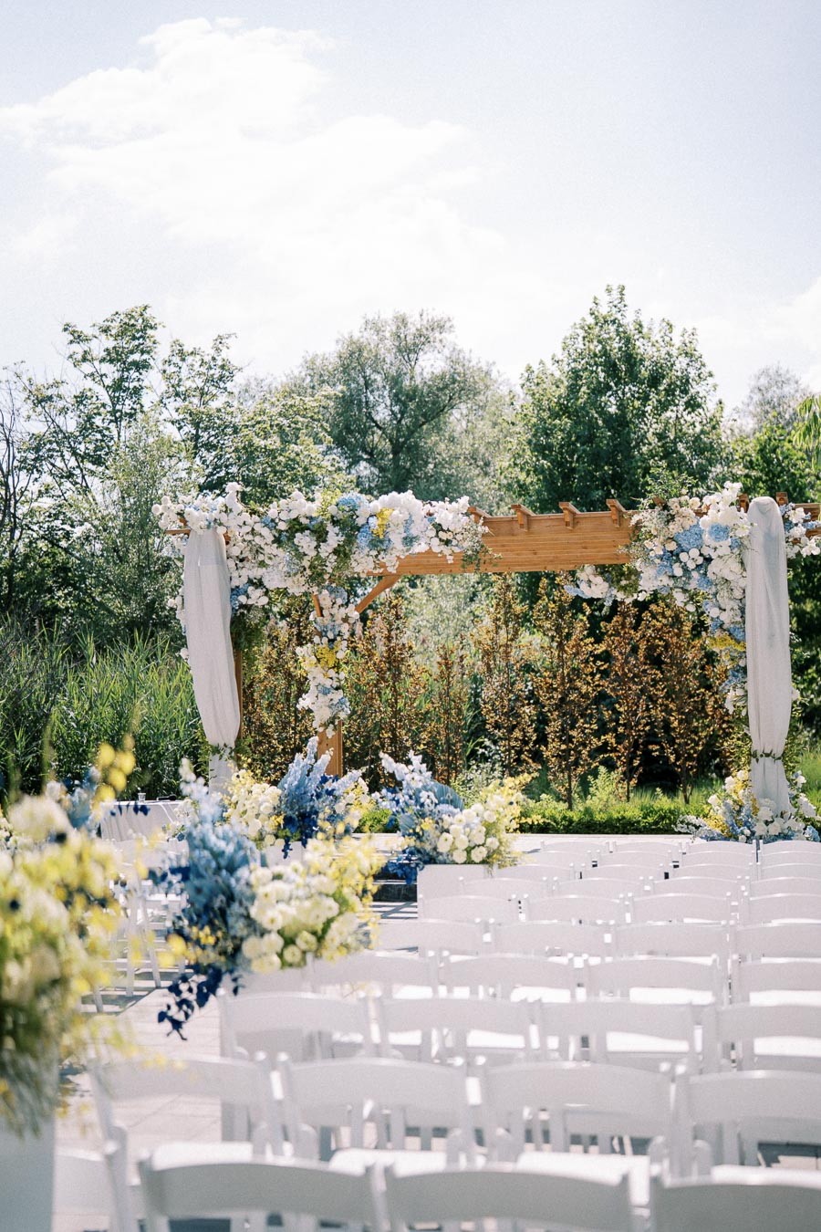 Outdoor wedding ceremony setup with white chairs and a wooden arch adorned with blue, white, and yellow flowers, set against a lush green garden backdrop under a clear sky.
