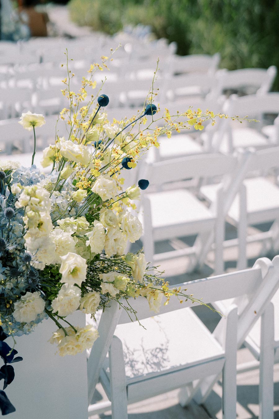White folding chairs arranged in rows for an outdoor wedding ceremony, adorned with a lush floral arrangement featuring yellow and blue blooms and greenery.