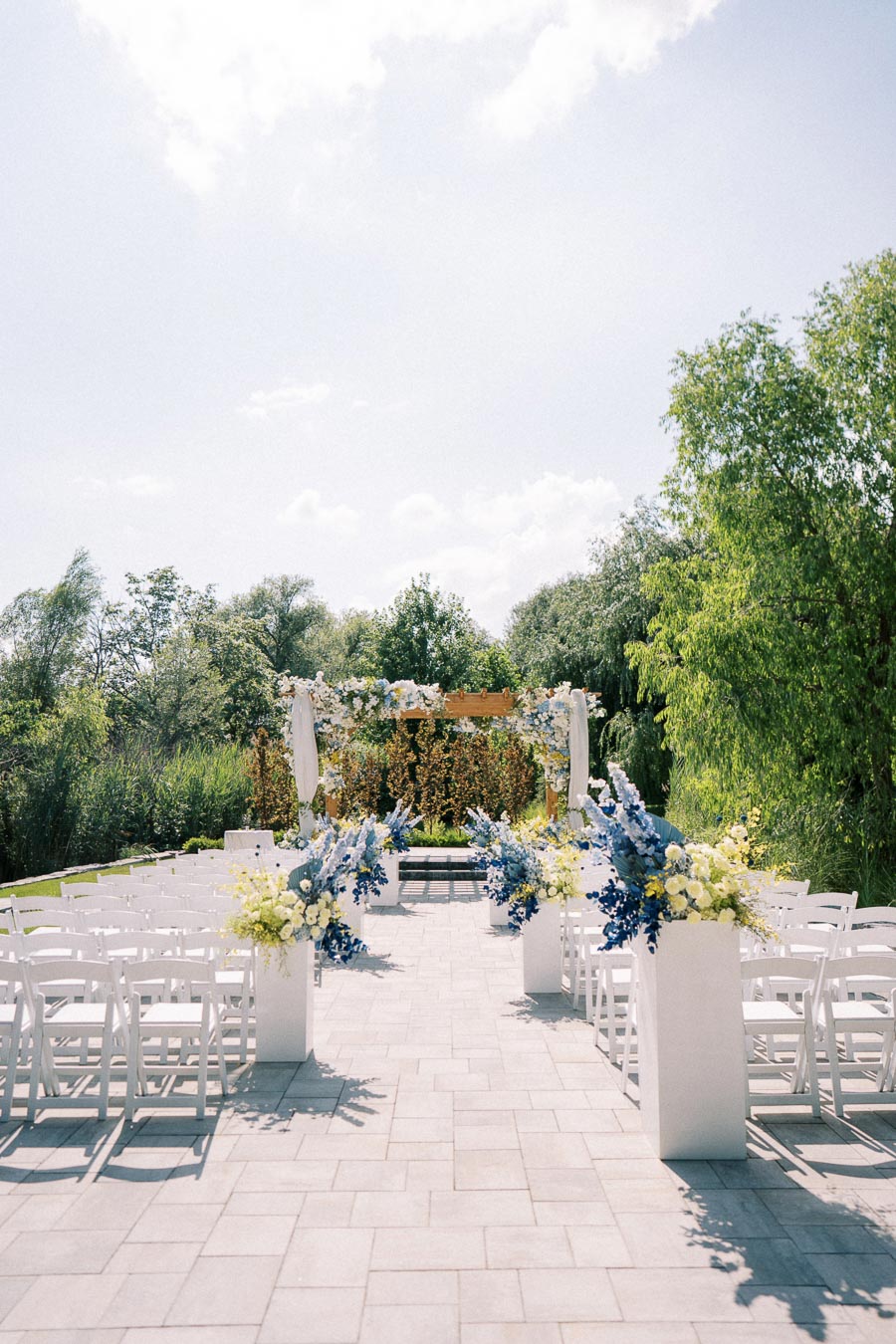 A beautifully decorated outdoor wedding ceremony setup with white chairs, a floral arch featuring blue and white flowers, and surrounding green trees under a clear sky.