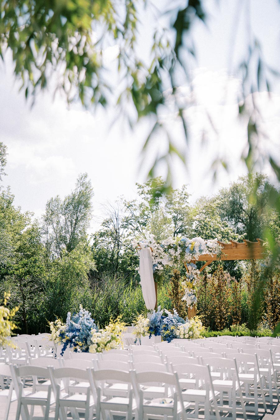 Outdoor wedding ceremony setup with white chairs and a floral-decorated wooden arch, surrounded by lush greenery and blue skies.