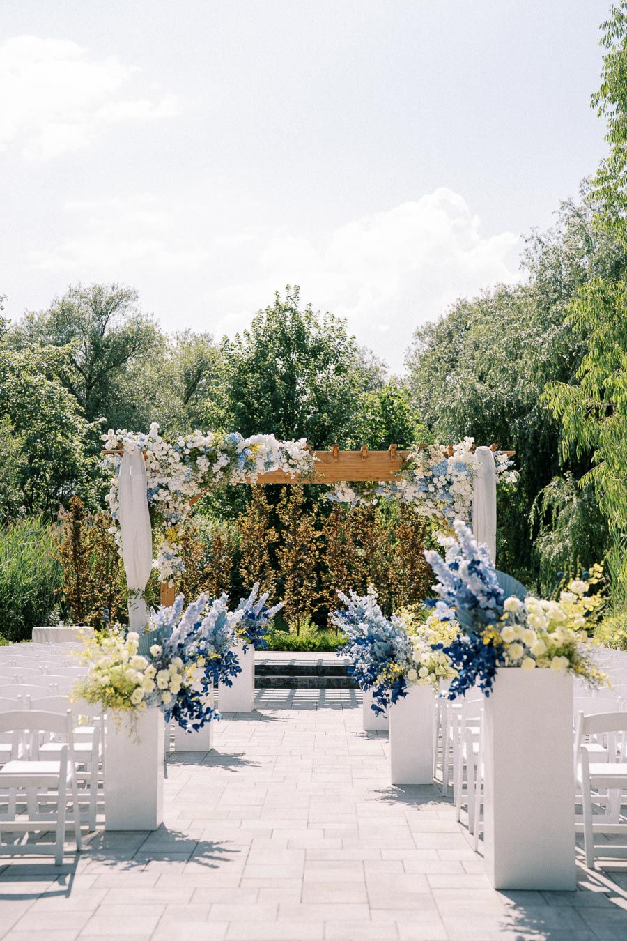 Outdoor wedding ceremony setting with a wooden arbor decorated with white and blue flowers, surrounded by lush green trees and white chairs lined up for guests. Bright and sunny day creating an elegant and serene atmosphere.