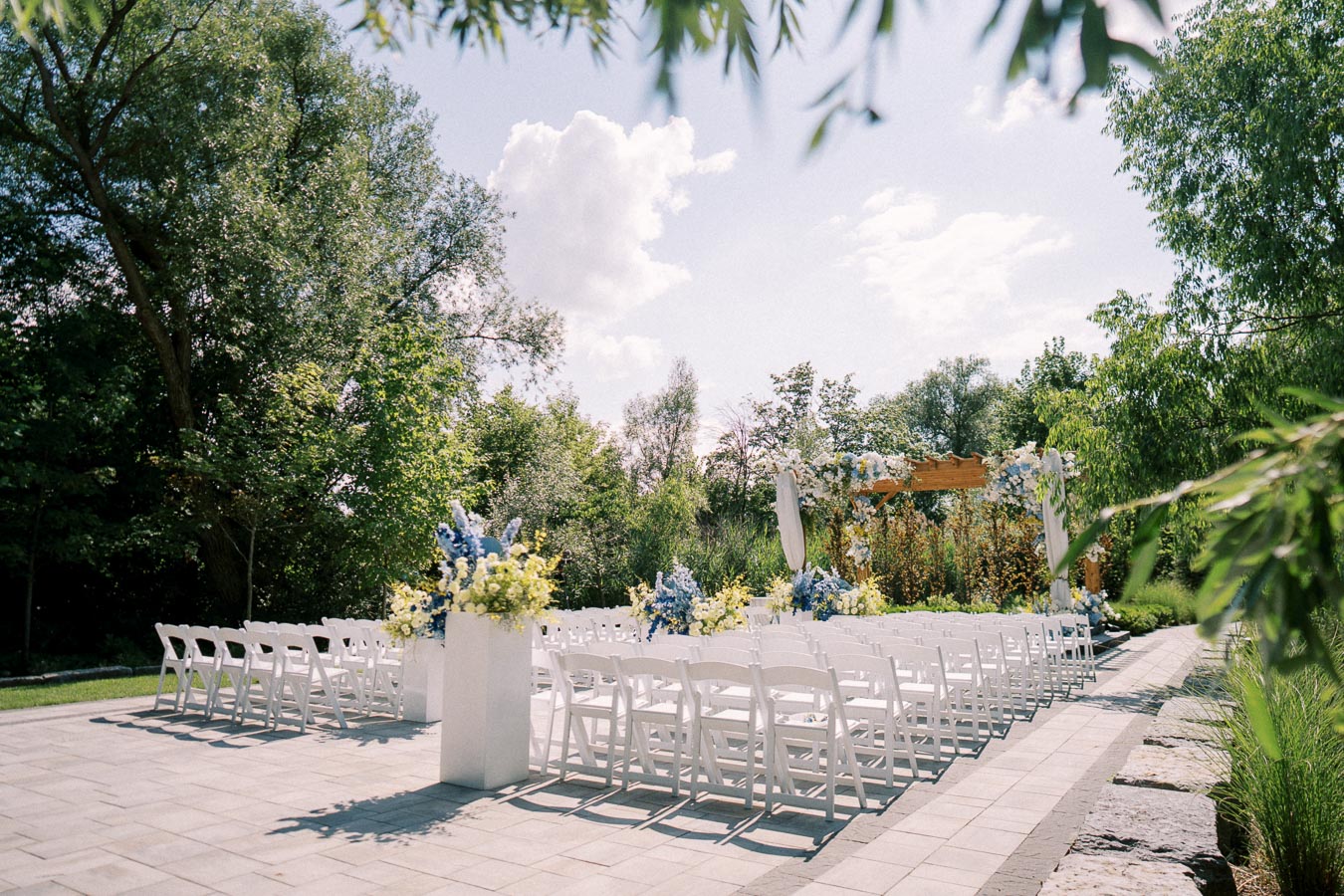 Outdoor wedding ceremony setup with white chairs and a floral-decorated archway, surrounded by lush greenery and under a bright blue sky.