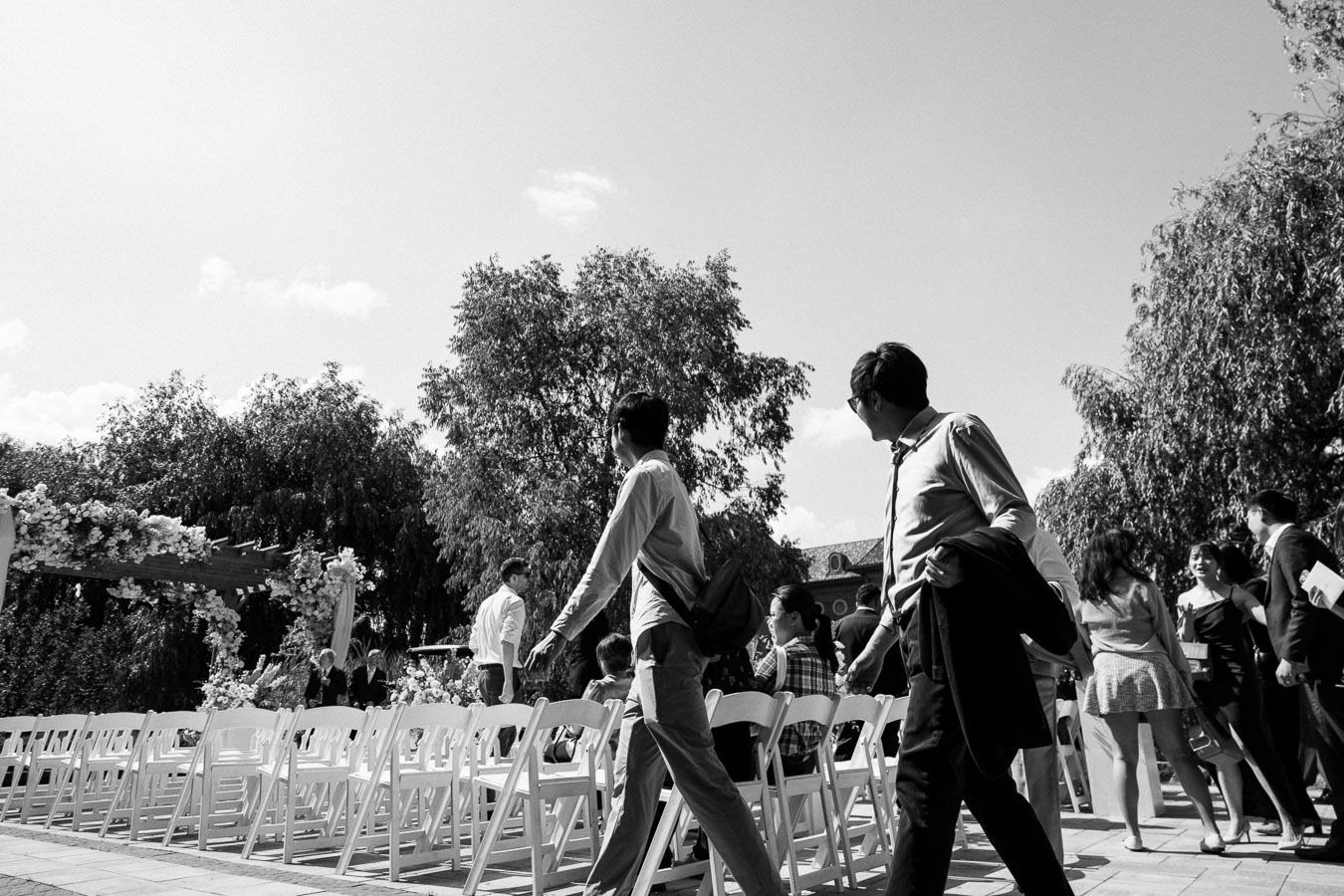 Black and white image of people walking around an outdoor wedding venue with white chairs and floral decorations under a clear sky.