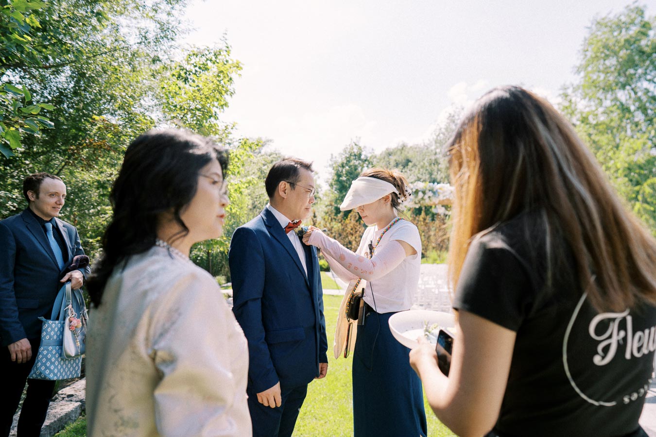 A woman pins a boutonniere on a man's suit jacket at an outdoor event, surrounded by people in formal attire, with lush greenery in the background.