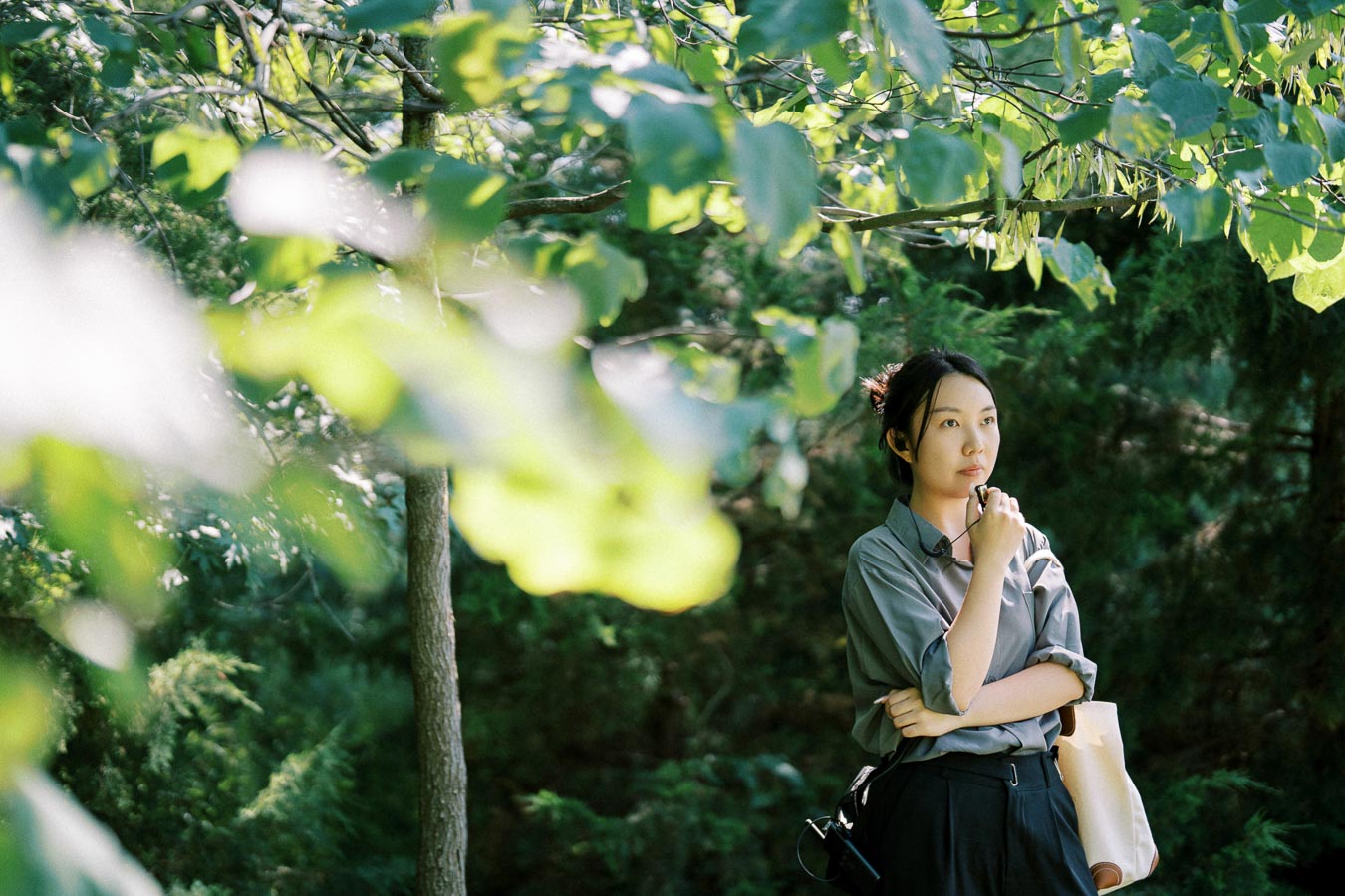 Young woman standing thoughtfully in a lush green forest, surrounded by sunlight-filtered leaves.