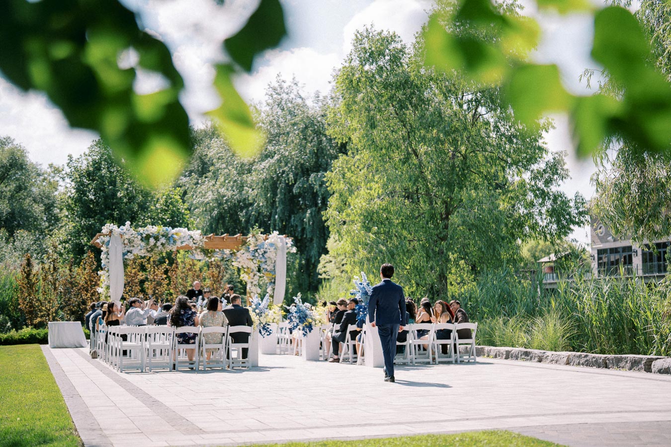 Outdoor wedding ceremony with guests seated and a groom walking down the aisle. The venue is surrounded by lush greenery and a floral arch, creating a picturesque setting on a sunny day.