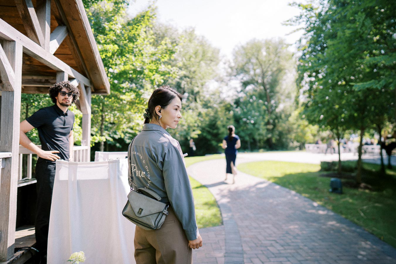 Outdoor scene featuring a woman in a casual jacket and crossbody bag walking on a pathway surrounded by lush green trees under a clear sky.