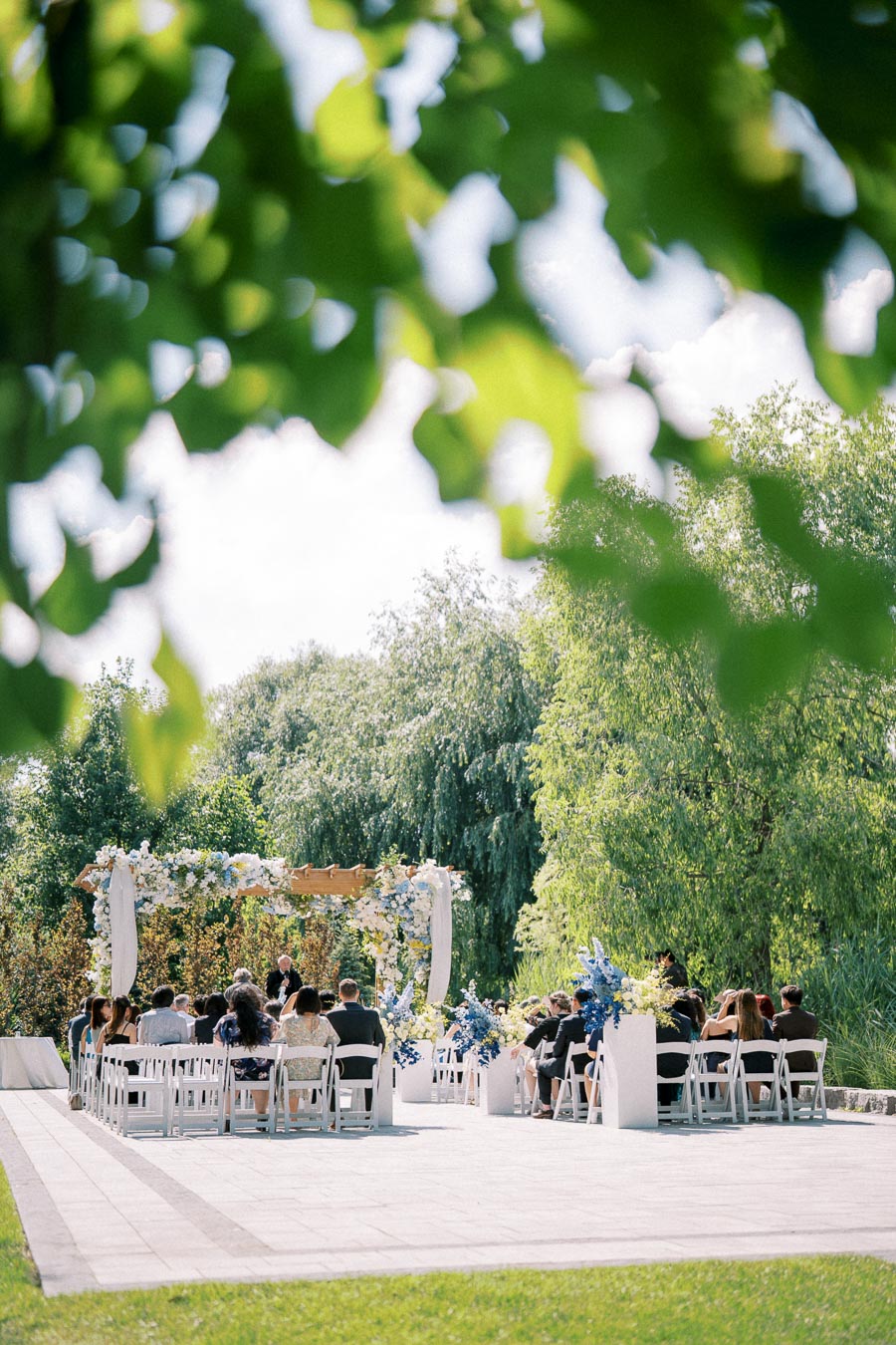 Outdoor wedding ceremony with guests seated in white chairs, surrounded by lush greenery and floral decorations.