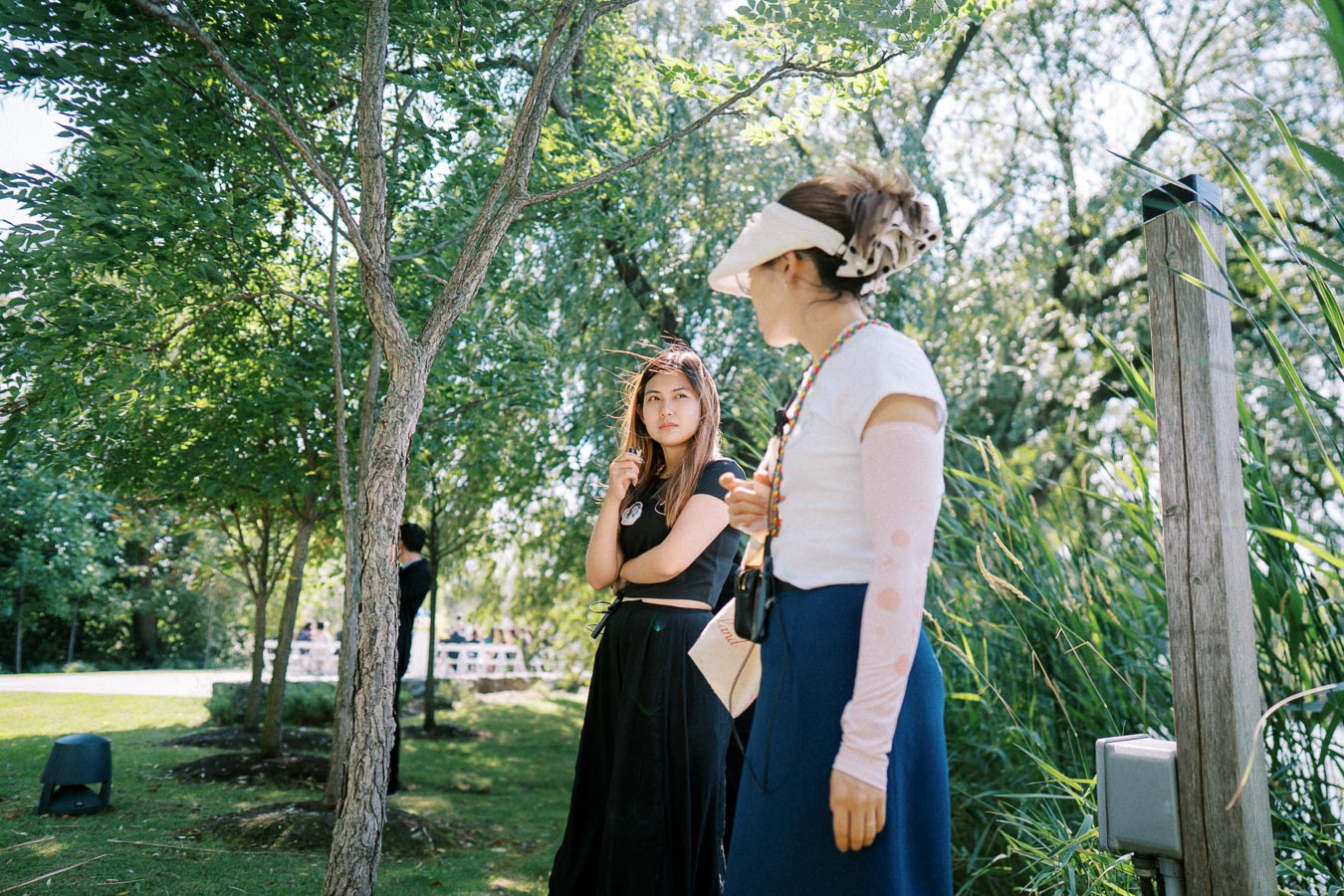 Two women standing in a sunlit park surrounded by lush green trees, engaging in conversation; one wearing a colorful beaded necklace and visor, the other in a black outfit, during a bright summer day.