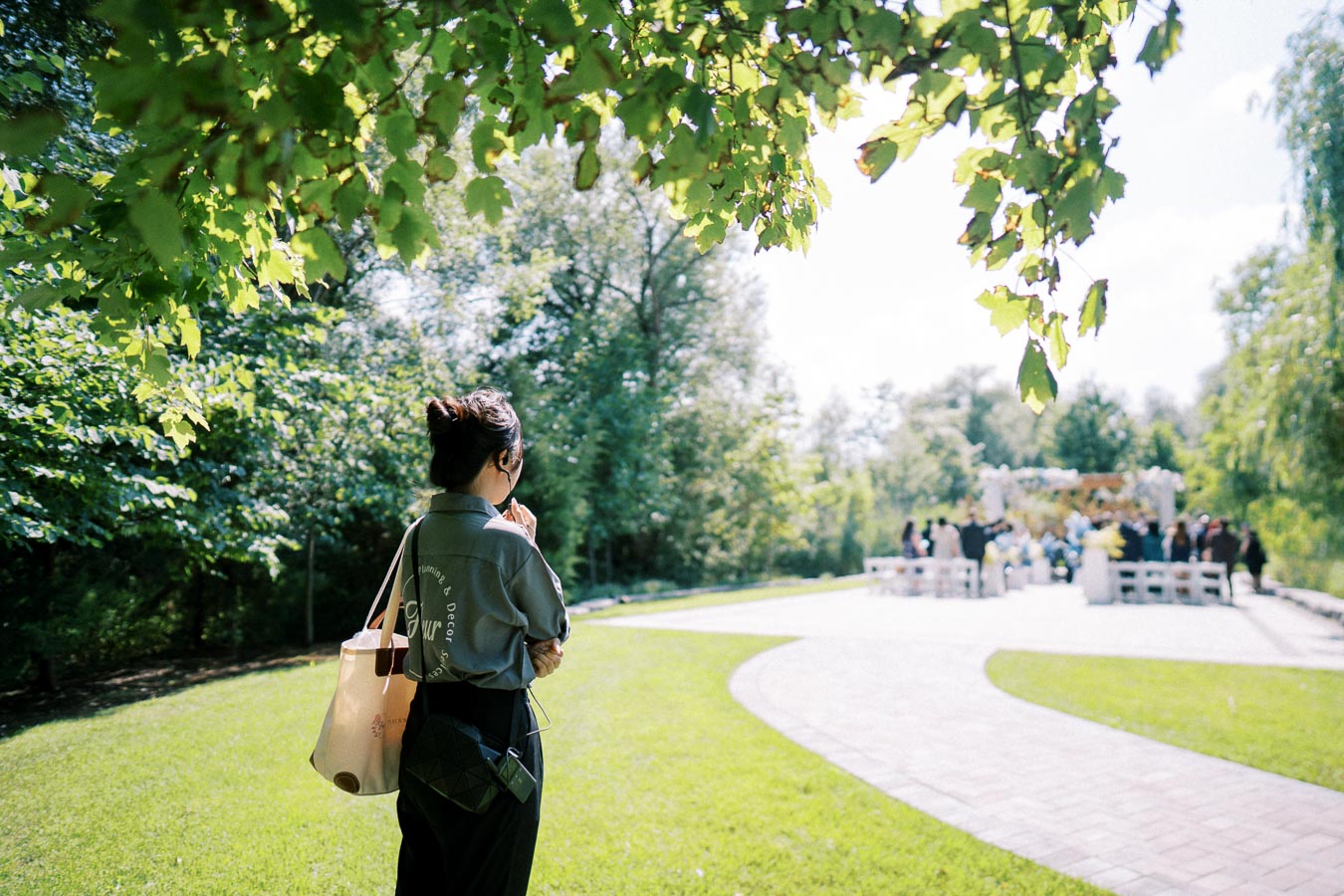 A woman stands in a sunlit park, observing an outdoor gathering with a path leading to an event area, surrounded by lush green trees and clear skies.